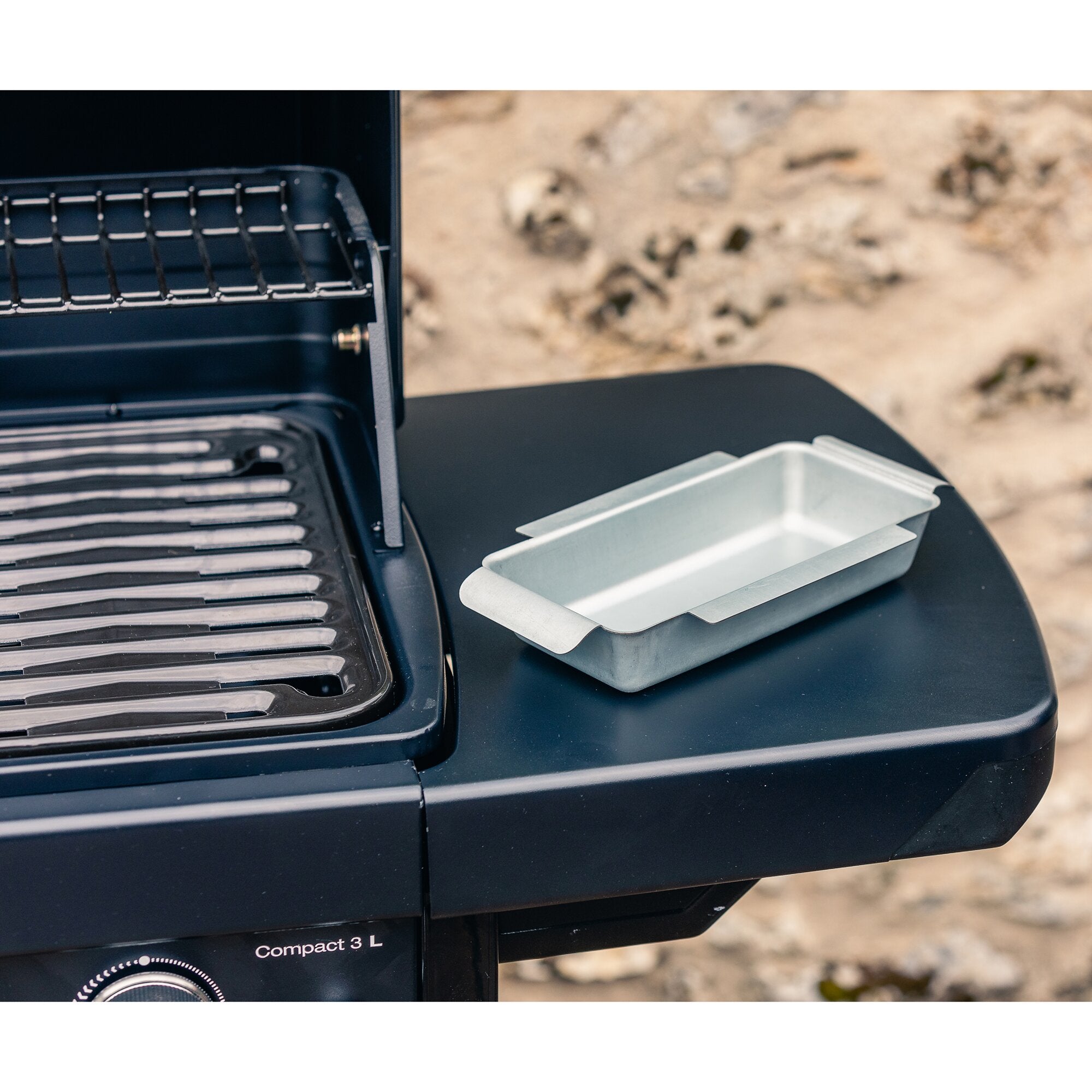 Teilansicht eines dunklen Gasgrills Compact 3 L: Innenraum mit Grillrost, rechts eine silberne Metallwanne mit mehreren Blechen auf der Ablage, unten Einstellknopf