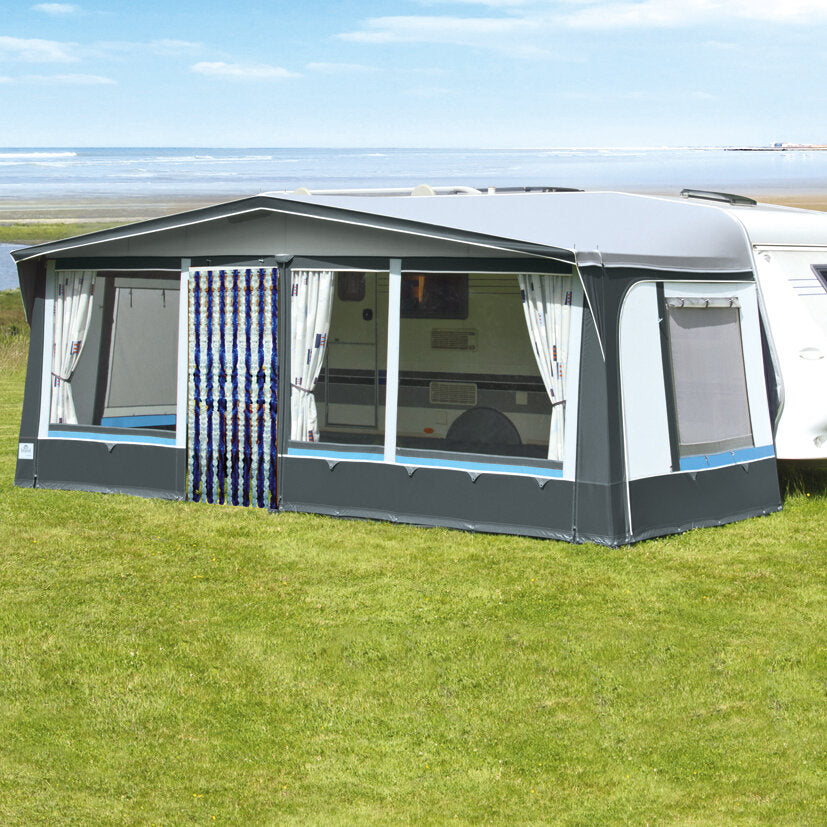 Caravan mit grauem Vorzelt und seitlichen Fenstern; weiße Vorhänge, blaue Leiste unten, grüne Wiese mit Strand und Meer im Hintergrund