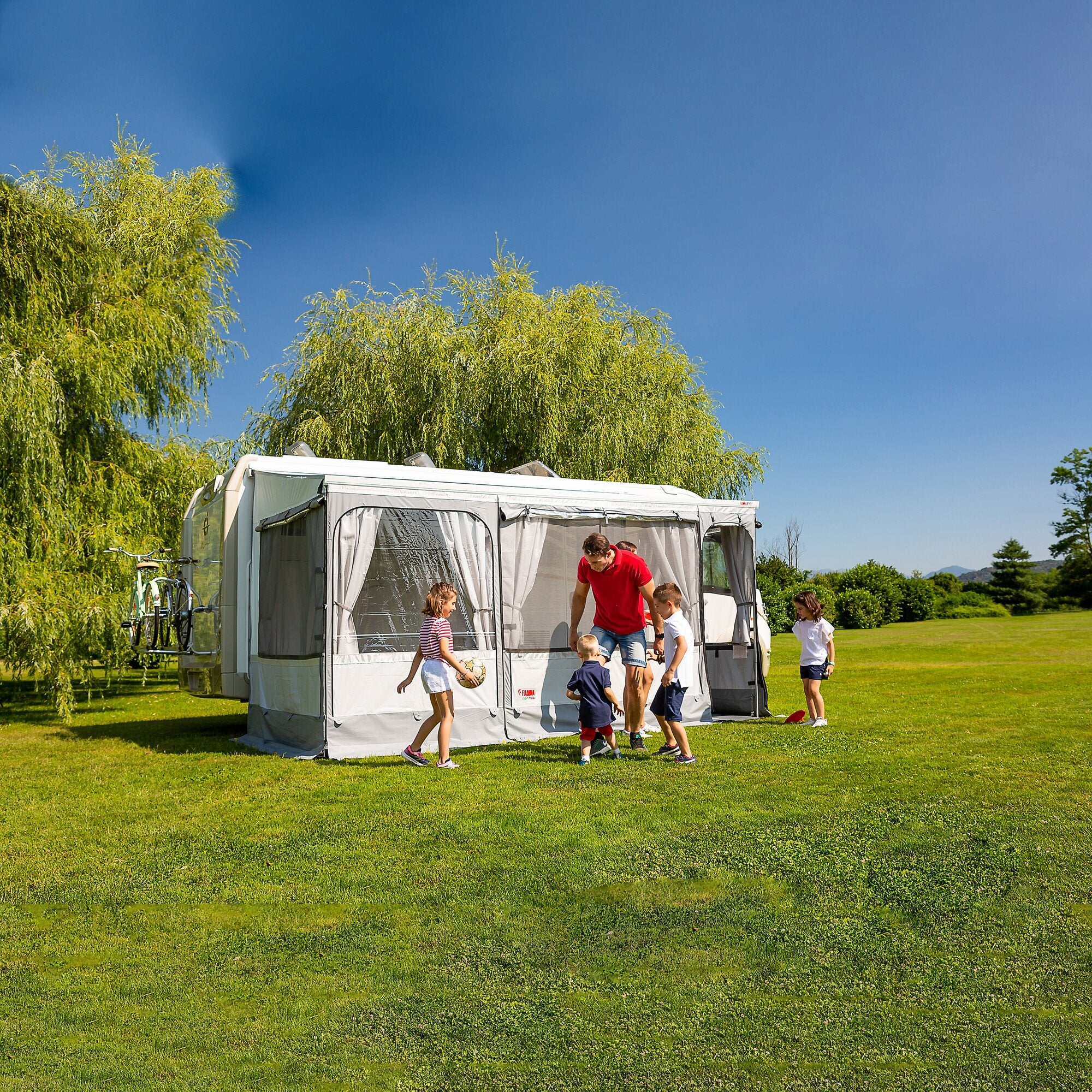 Campingplatz mit weißem Wohnwagen und Markisenzelt Privacy Room F45, 300 Large; Kinder spielen Ball vor der Zeltfront, Mann hilft, Hintergrund Bäume