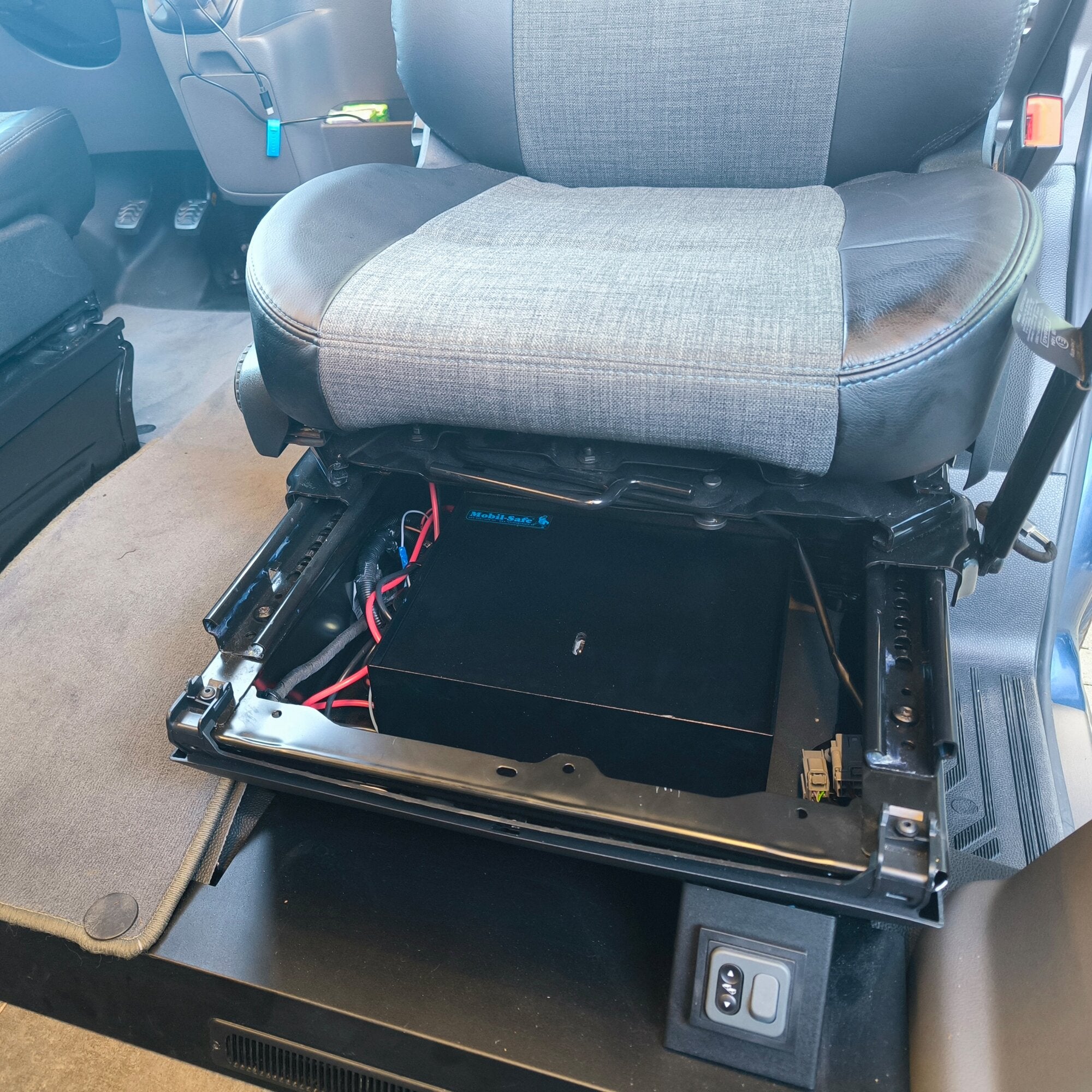 Sitzsockelsafe unter dem Fahrersitz eines Ford Transit ab 2019; grauer Stoffbezug, schwarzer Metallrahmen und sichtbare Kabel
