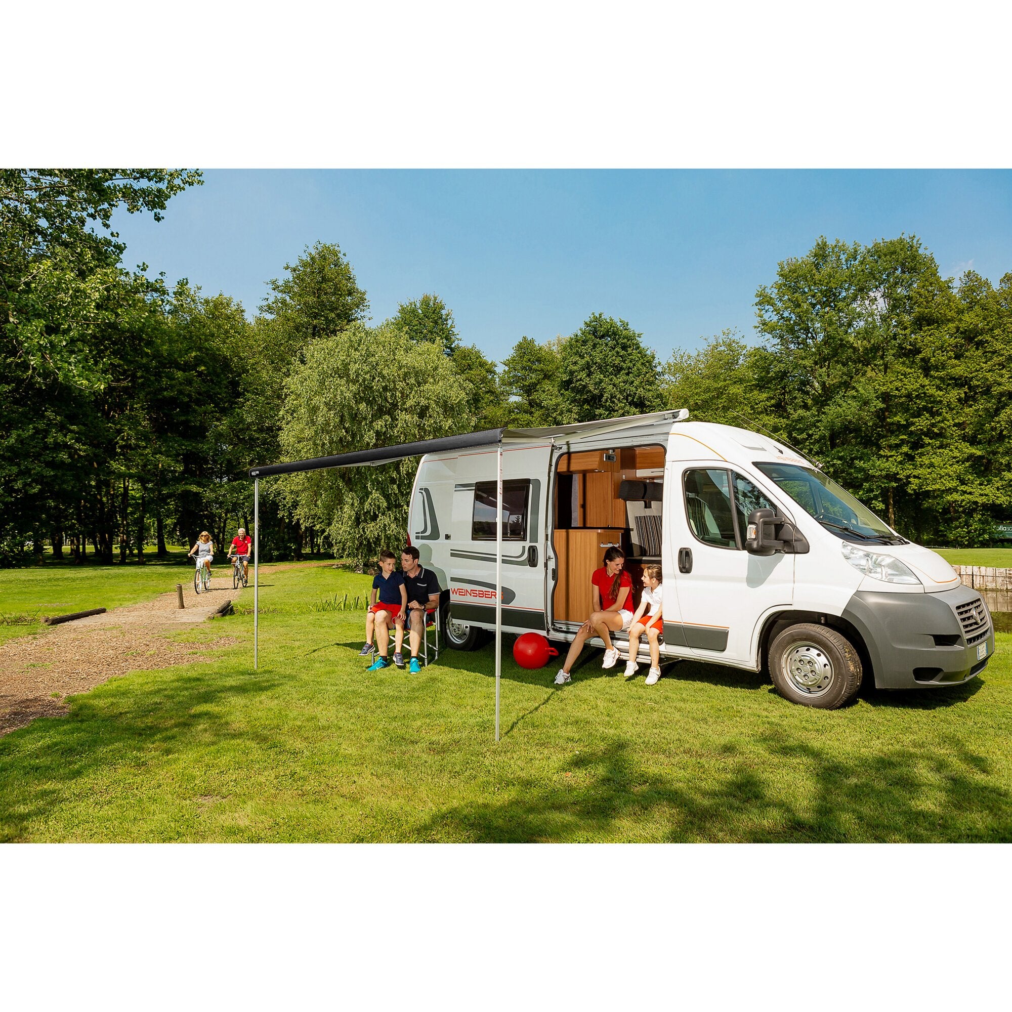 Schwarze Markise F80 am weißen Wohnmobil ausgefahren, davor sitzen mehrere Personen im Park, grüne Bäume und blauer Himmel im Hintergrund