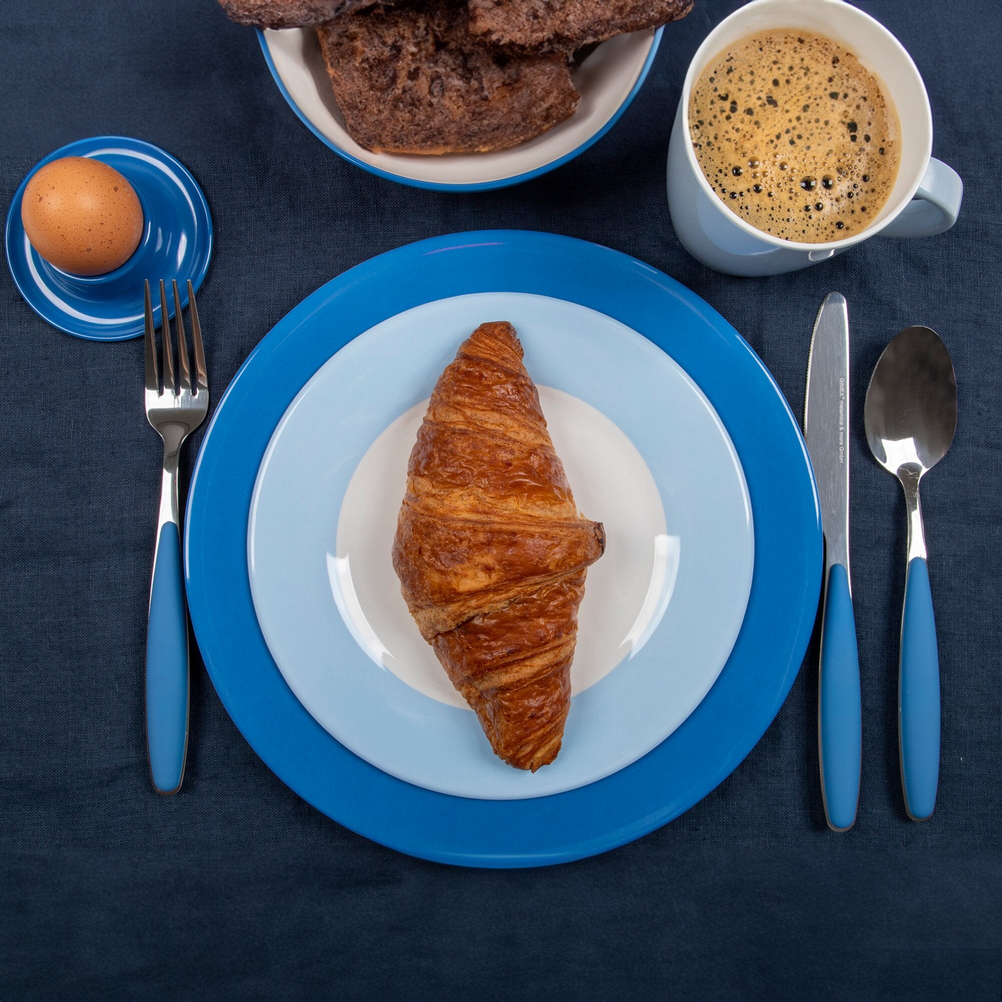 Top-Ansicht eines blauen Colour Line Geschirrsets auf dunkelblauem Tisch: Teller, Croissant, Kaffeetasse, Ei im Eierbecher, Besteck