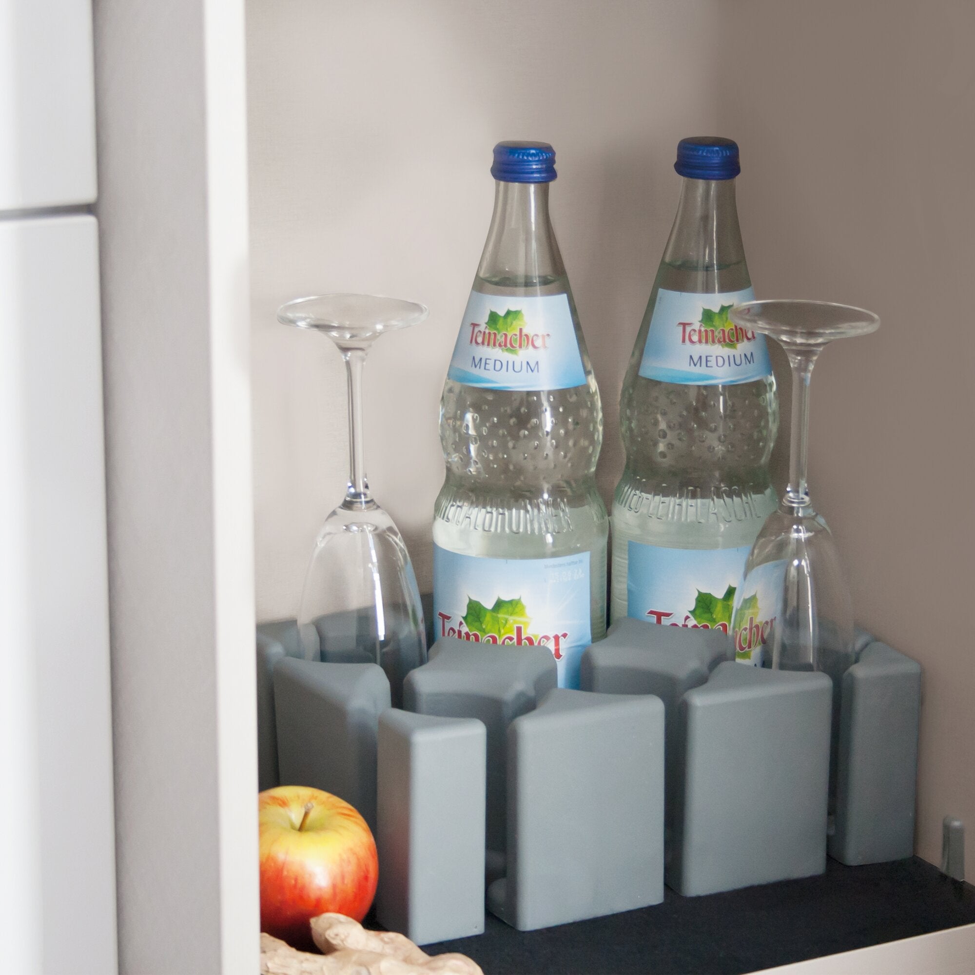 Offener Küchenschrank mit zwei Teinacher Medium Mineralwasserflaschen, Deckeln, zwei Weingläsern, Äpfeln und Ingwer vor grauen Trennwänden