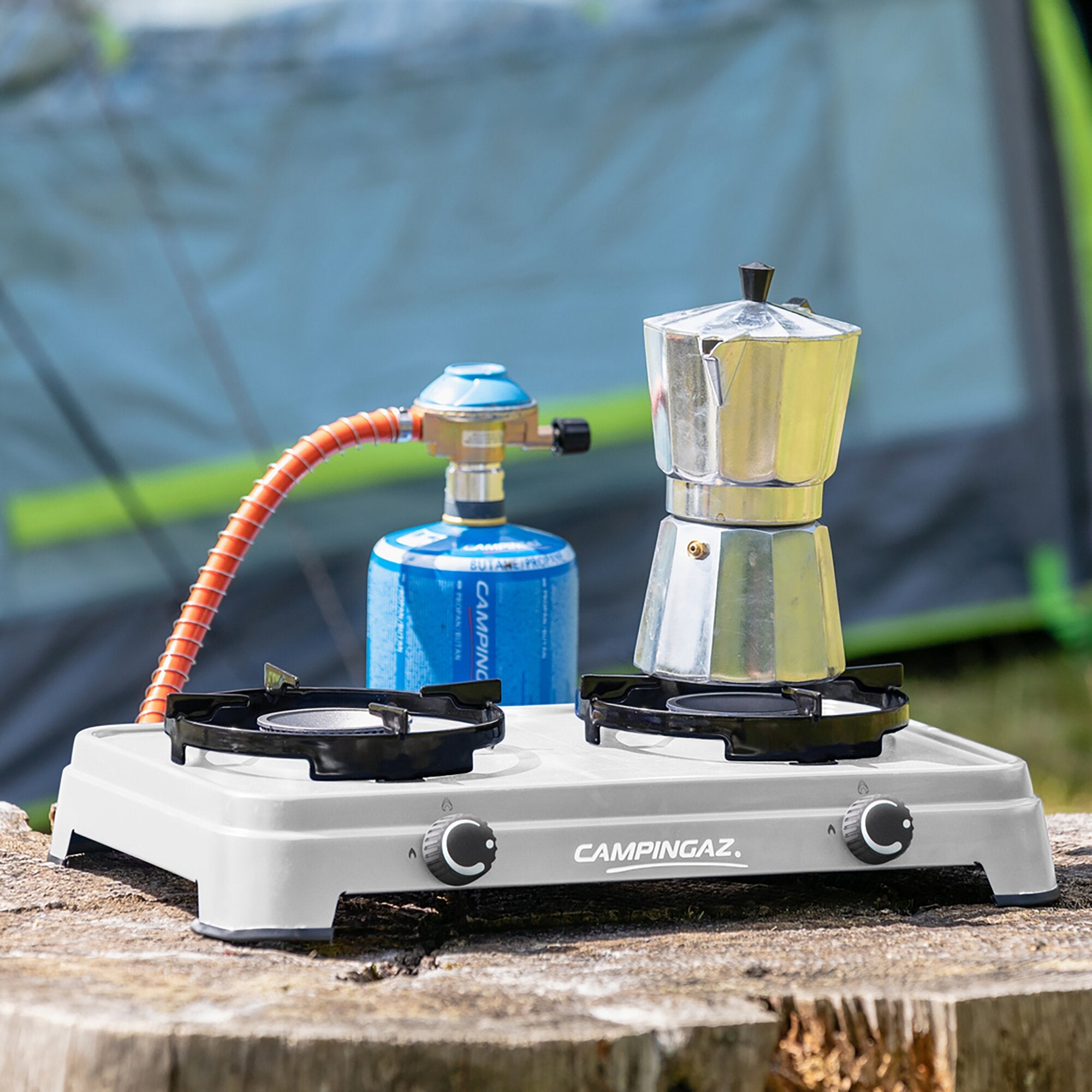 Campingkocher mit zwei Brennern, silberne Moka-Pot auf dem Herd, orange Gasschlauch zum blauen Campingaz-Gasbehälter, unscharfes Zelt im Hintergrund