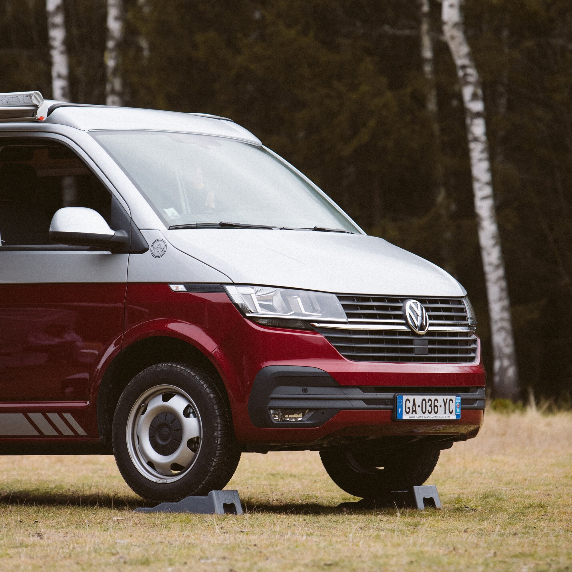 Zwei graue Auffahrkeile Level Up 2er-Set liegen unter dem vorderen Rad eines weißen Vans, Wald im Hintergrund
