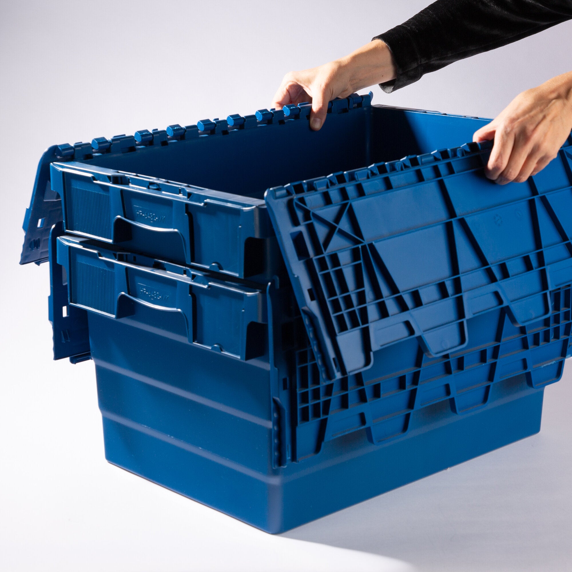Nahaufnahme einer blauen Kunststoff-Transportbox mit Oberkante aus Verriegelungselementen; eine Hand greift hinein, dahinter stapeln sich weitere Boxen