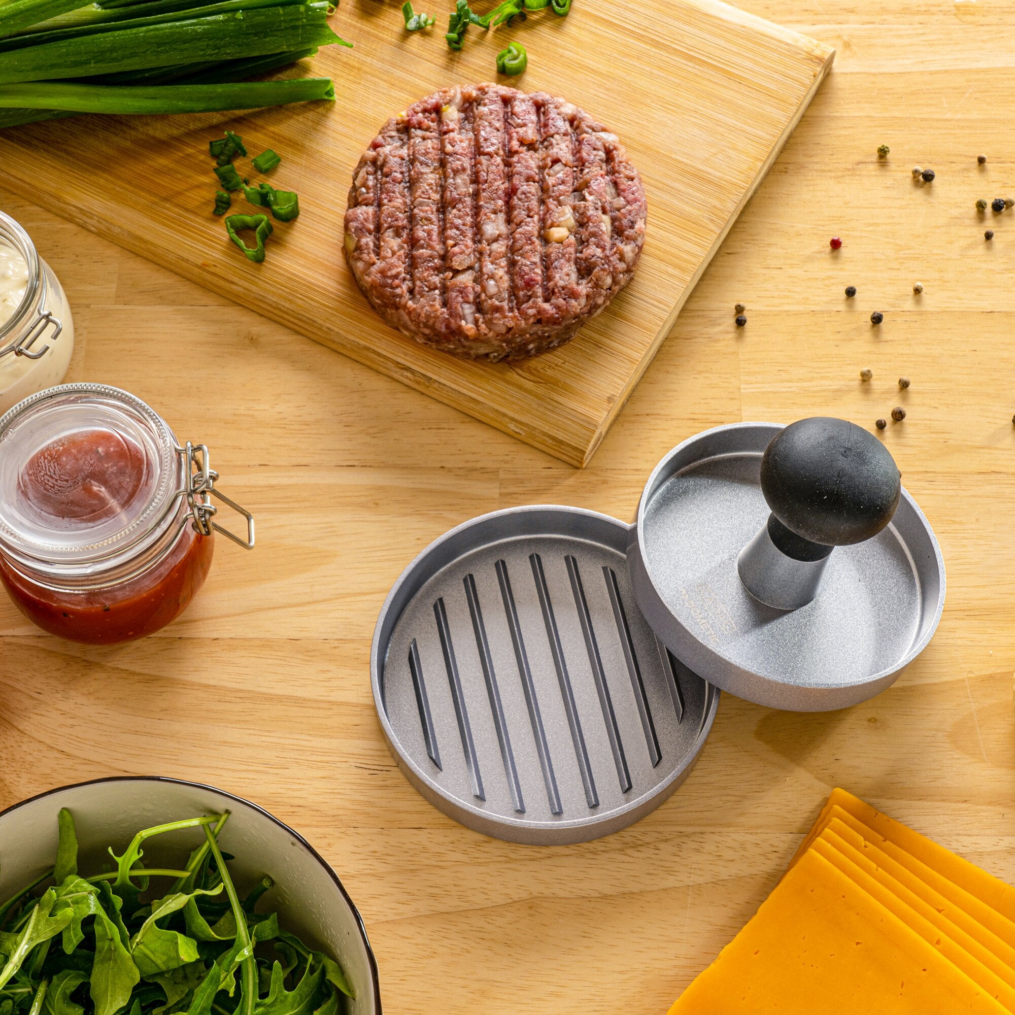 Runde Hamburgerpresse aus Metall mit Deckel neben einem geformten Hackfleischpatty auf einem Holzbrett, umgeben von Käse und Salatblättern