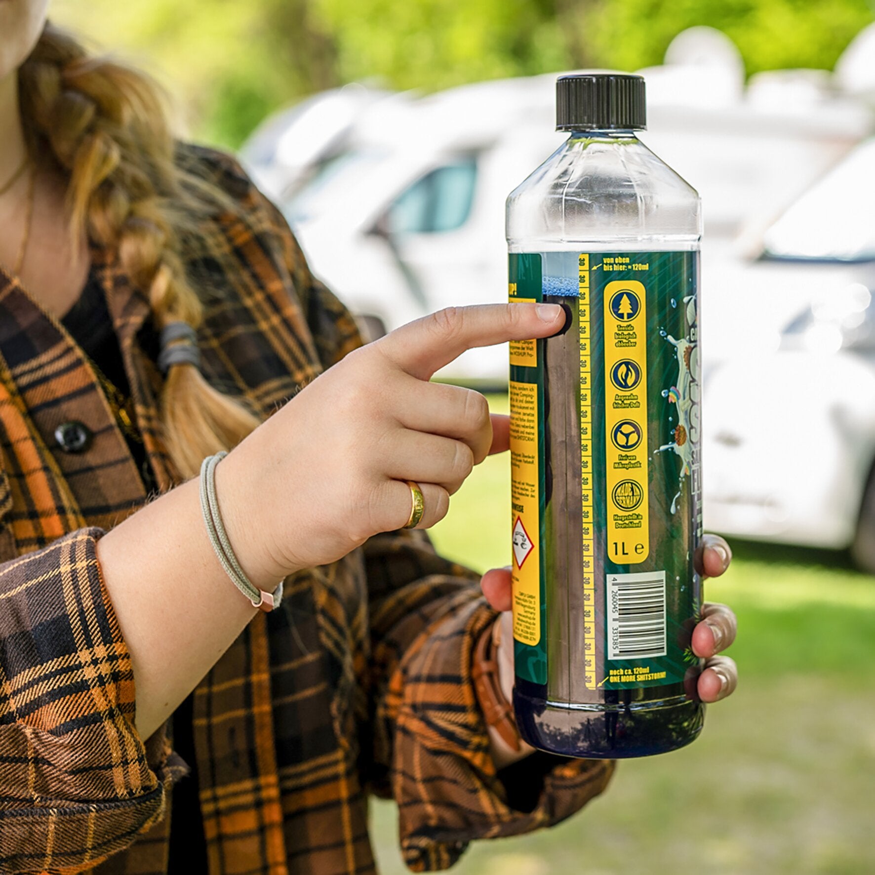 Person in kariertem Flanellhemd hält eine 1-L-Flasche Shitstorm Tanne Toilettenzusatz, grün-gelbes Etikett mit Baum-Symbol, schwarzer Deckel, im Freien