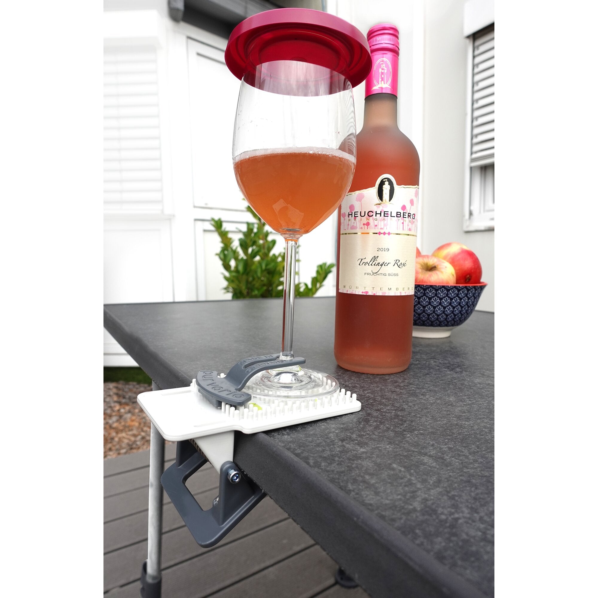 Trinkglasabdeckung BEE sitzt auf einem Weinglas mit orangefarbenem Getränk; im Hintergrund eine rosa Weinflasche und Obst auf der Terrasse