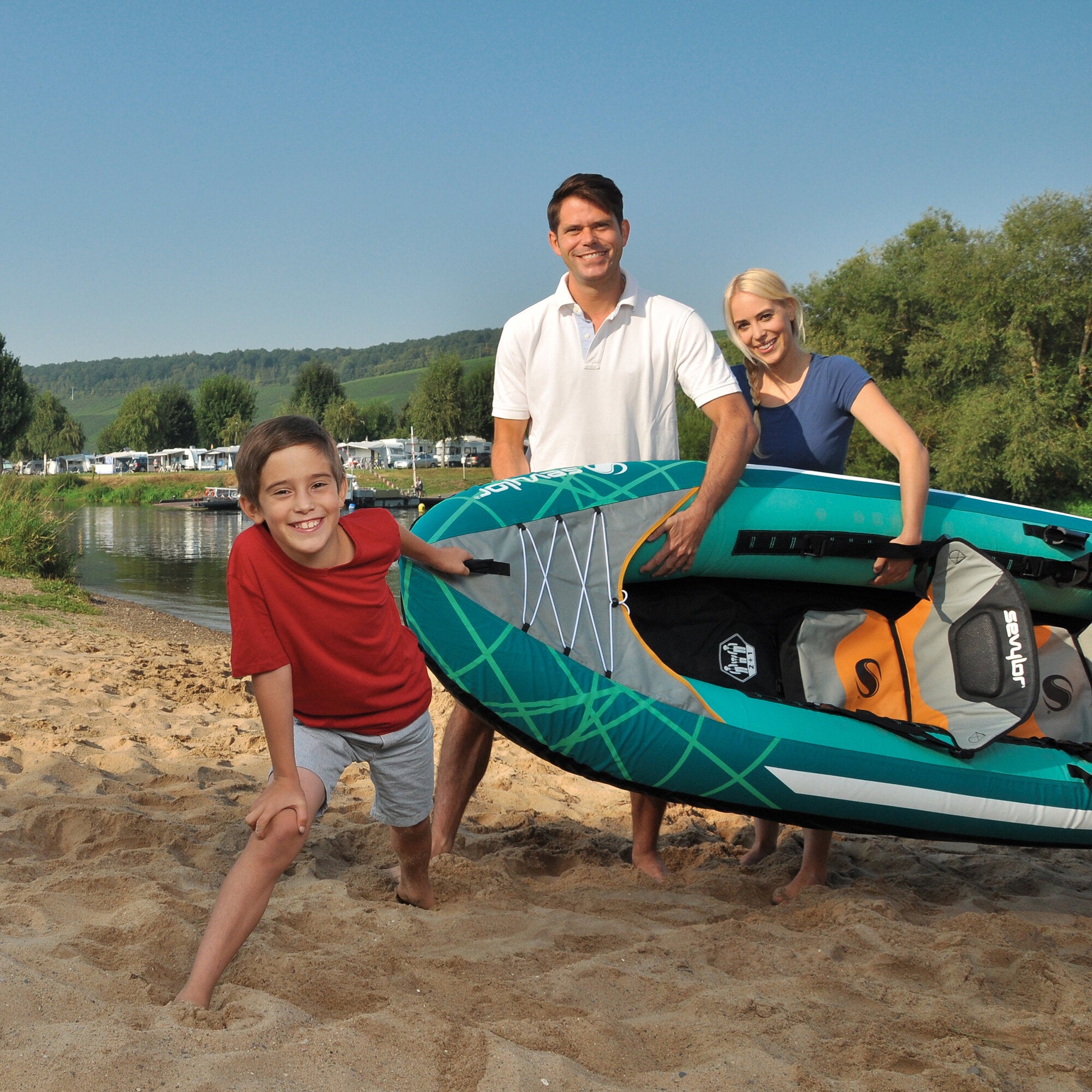 Familie aus zwei Erwachsenen und Kind am Strand hält ein aufblasbares Kajak Alameda in türkis mit orangefarbenen Akzenten; Hintergrund Wald, Wasser und Zelte