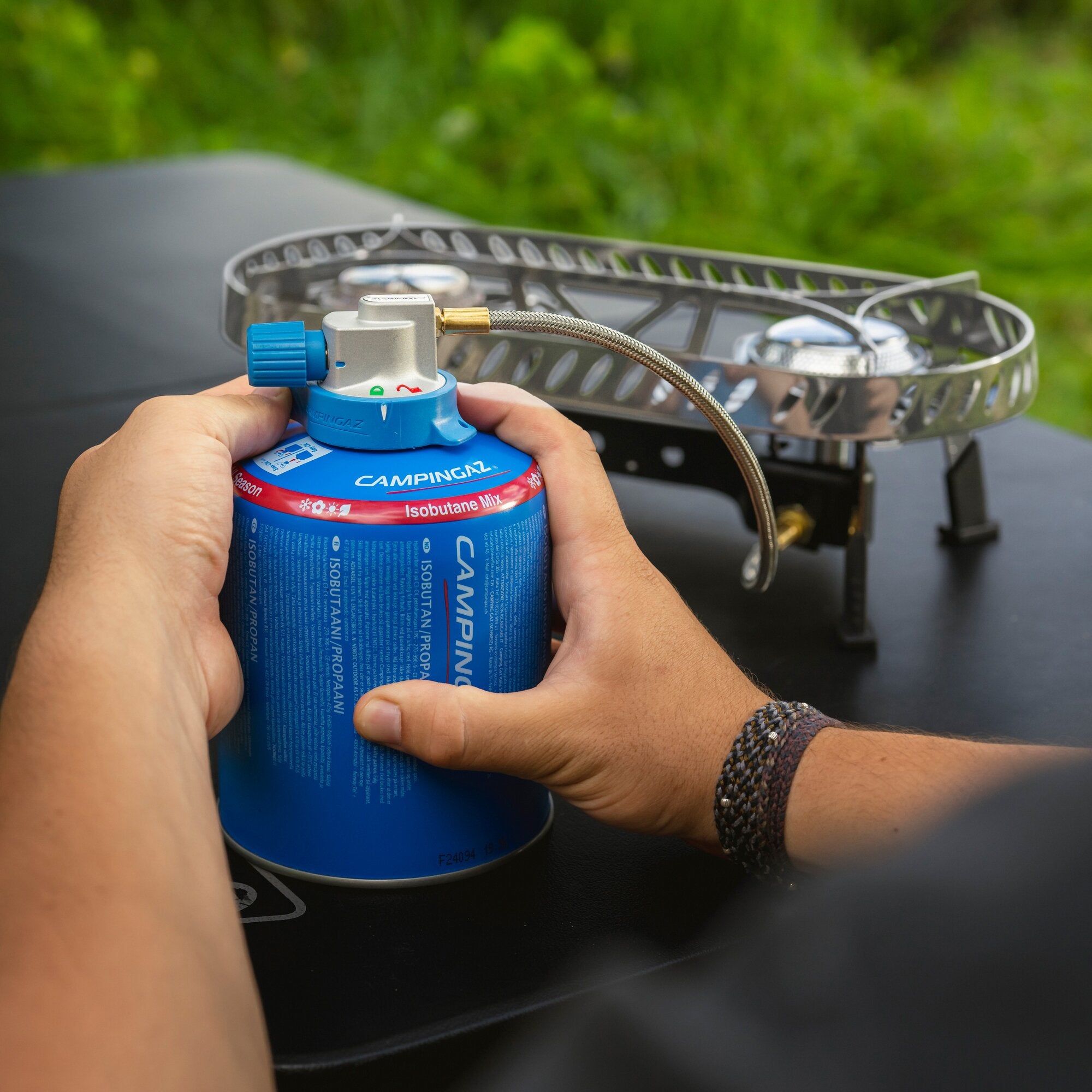 Nahaufnahme einer blauen Campingaz Gasflasche mit Regler und Edelstahl-Schlauch, der an einen tragbaren Campingkocher angeschlossen ist, grüner Hintergrund unscharf