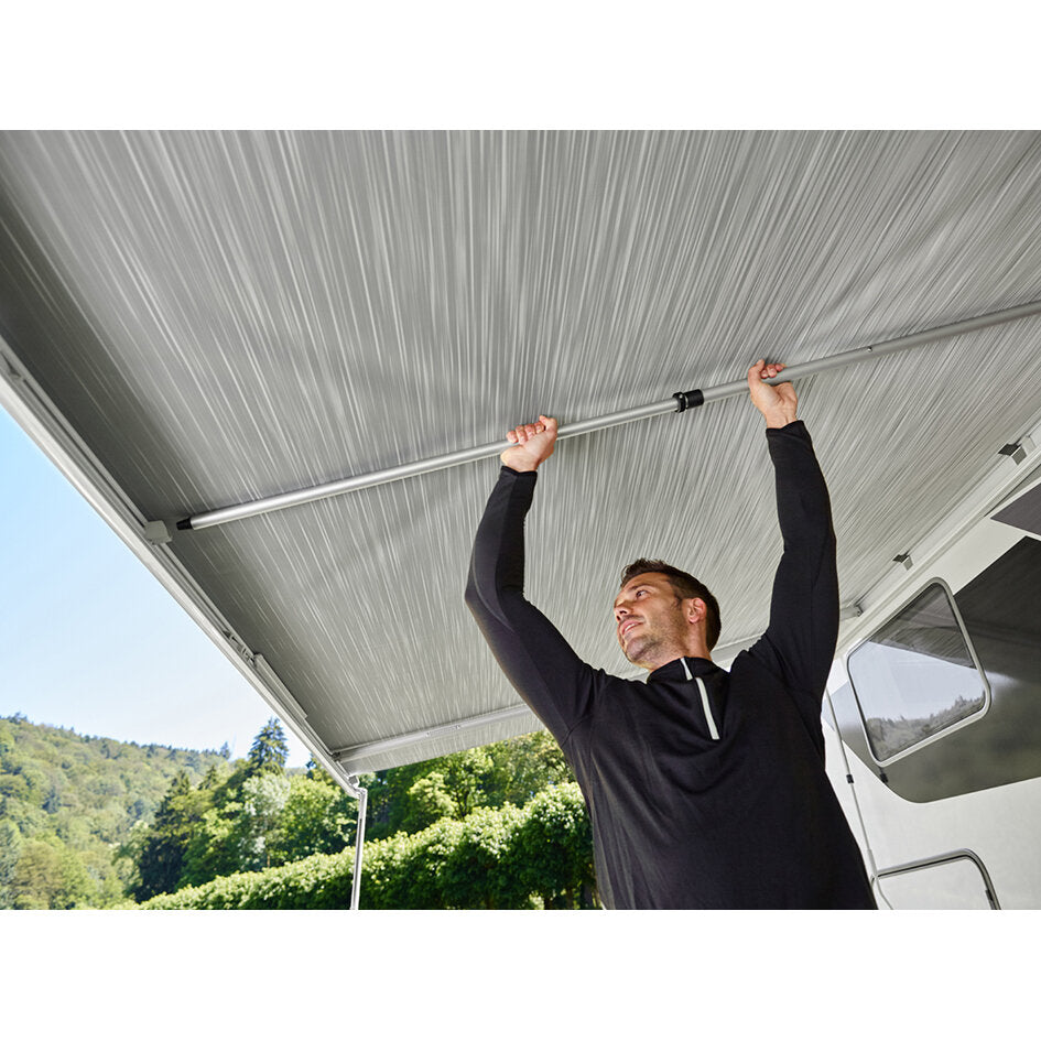 Ein Mann hält eine lange Metallspannstange (2 m) horizontal unter der ausfahrbaren Wandmarkise eines Wohnmobils, draußen vor grüner Landschaft