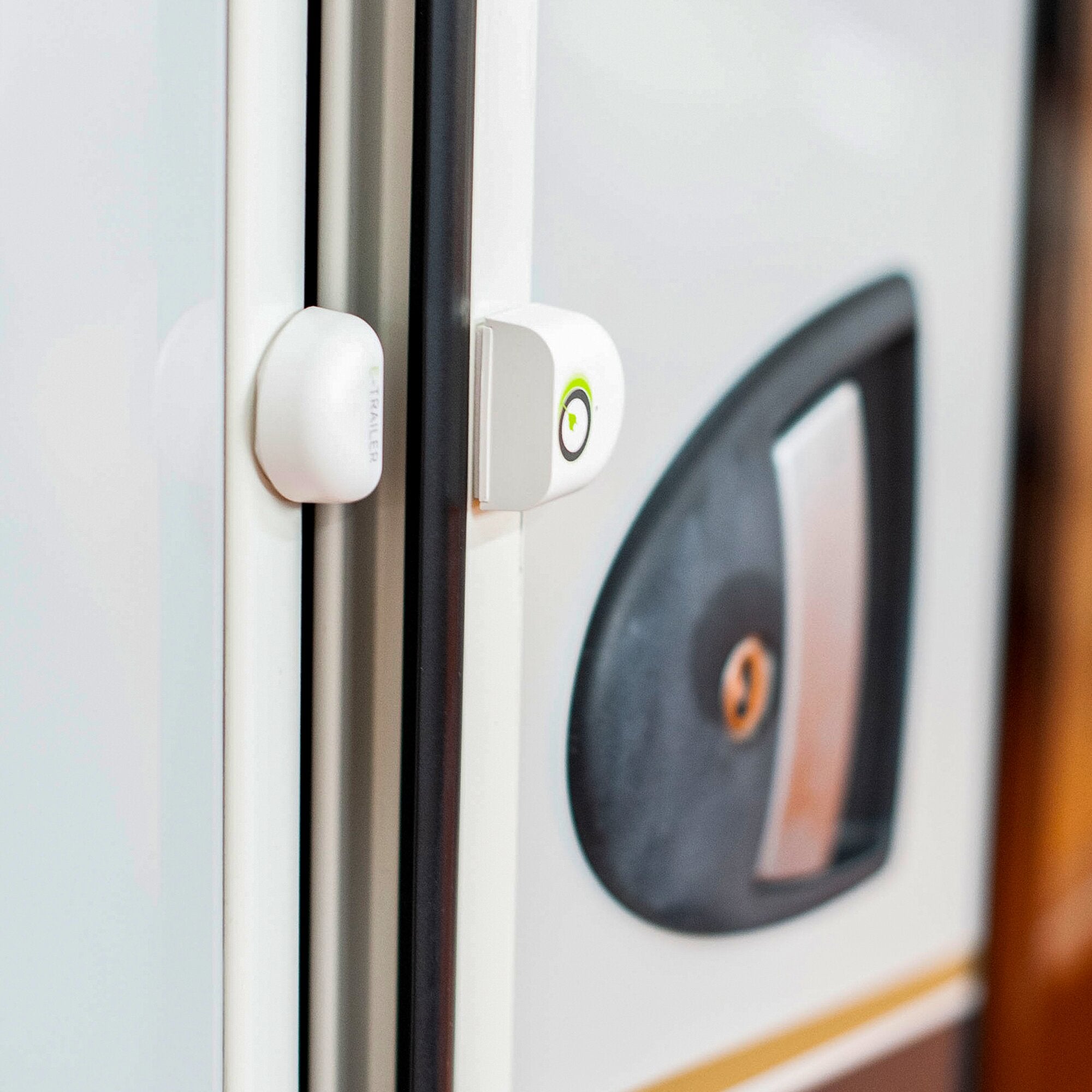 Nahaufnahme eines weißen E-Switch Tür-/Fenster-Sensors am Türrahmen, Gehäuse mit grünem Akzent, Glasfenster im Hintergrund