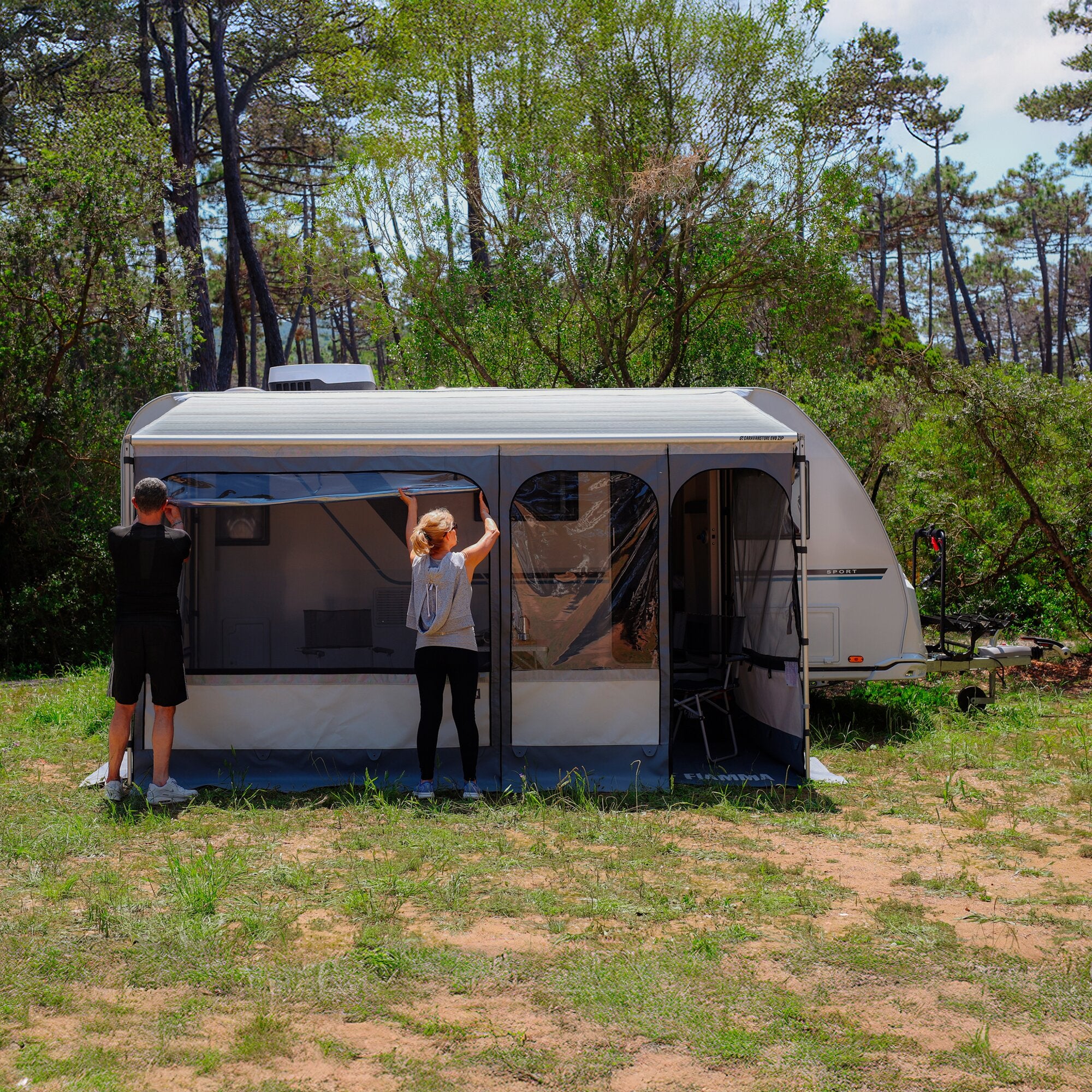 Caravan mit ausgefahrener Markise CaravanStore EVO ZIP 320 Royal Grey; zwei Personen stehen davor und richten die Vorzeltwand aus, Wald im Hintergrund