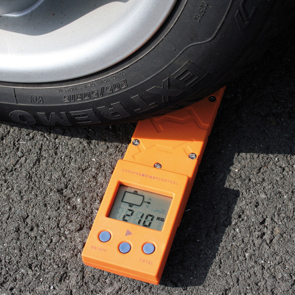 Orangefarbene Stützlastwaage Caravan Weight Control mit digitalem Display zeigt 210 kg liegt neben einem Reifen auf Asphalt