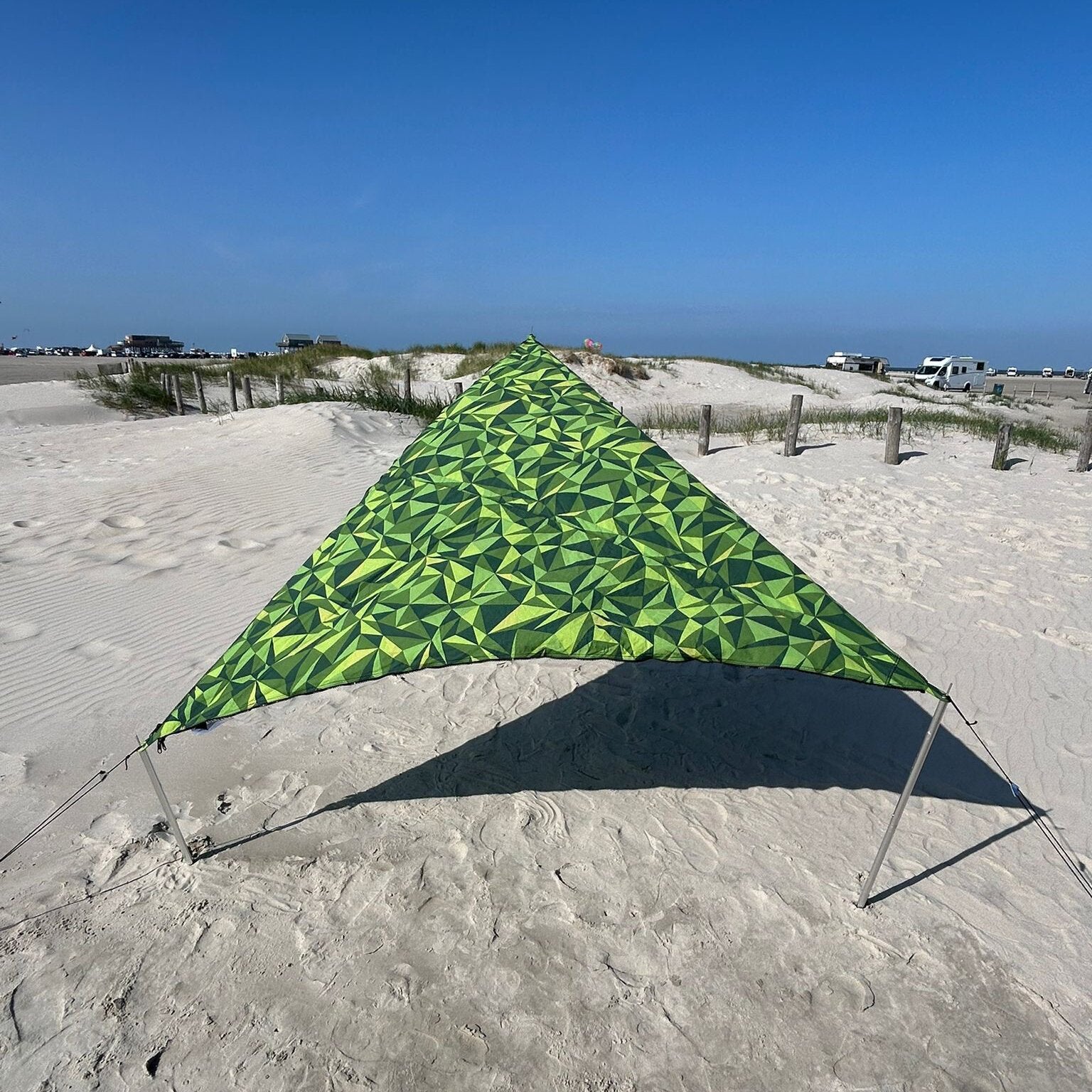 Grünes sonnensegel mit geometrischem Muster aus Dreiecken, am Sandstrand gespannt, Seile und Stangen befestigen das Segel, Dünen und Autos im Hintergrund