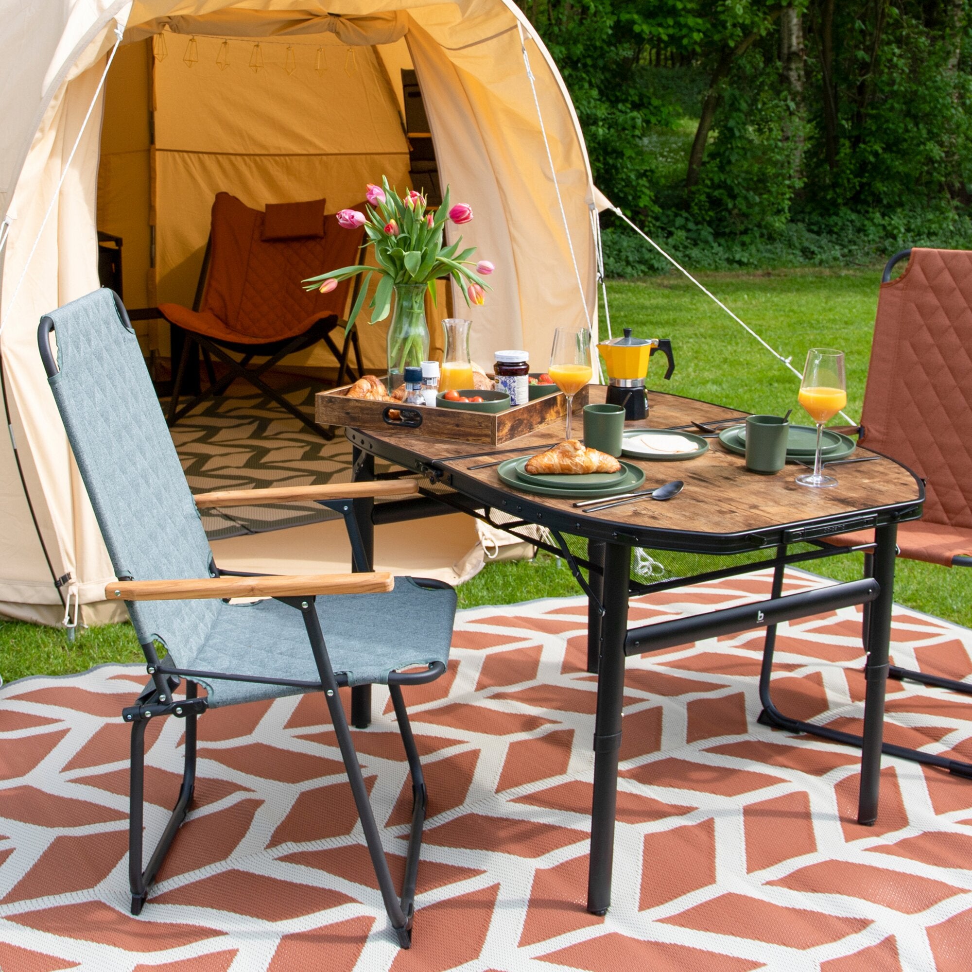 Campingtisch Woodbine 150 x 80 cm, Holzoptik-Tischplatte, Gestell aus Metall, im Freien gedeckt mit Tulpen, Croissants und Saft vor Zelt
