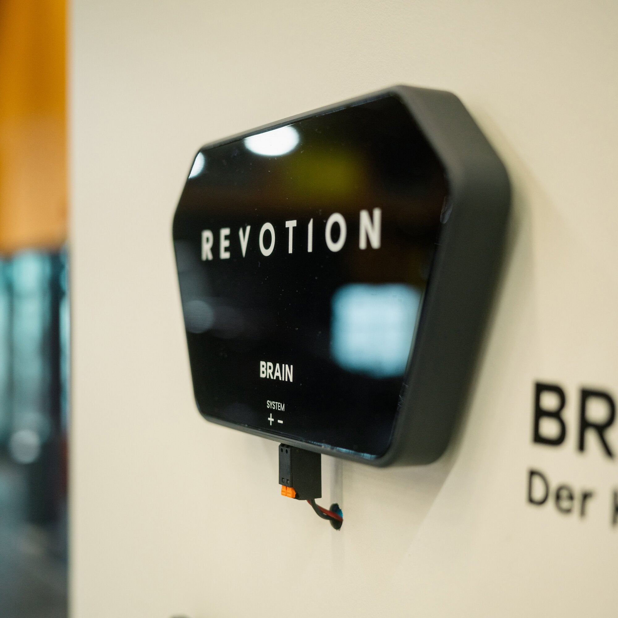 Schwarzes, glänzendes Gehäuse mit der Aufschrift REVOLUTION BRAIN; unten Kabel mit orangefarbenem Stecker, montiert an einer Wandplatte