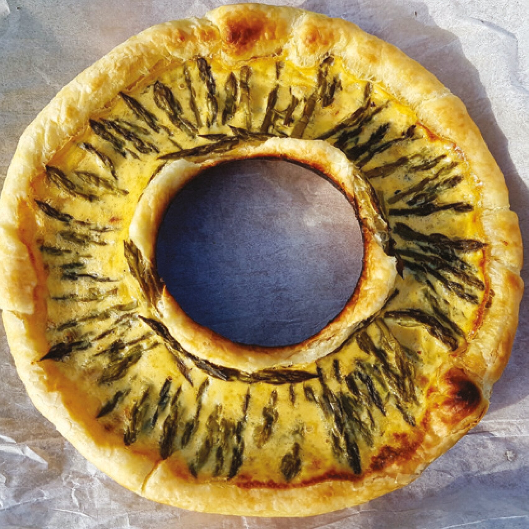 Runde Quiche in Ringform mit goldbrauner Kruste und gelbem Teig, verziert mit dunklen Kräuterstreifen rund ums Loch, auf Backpapier