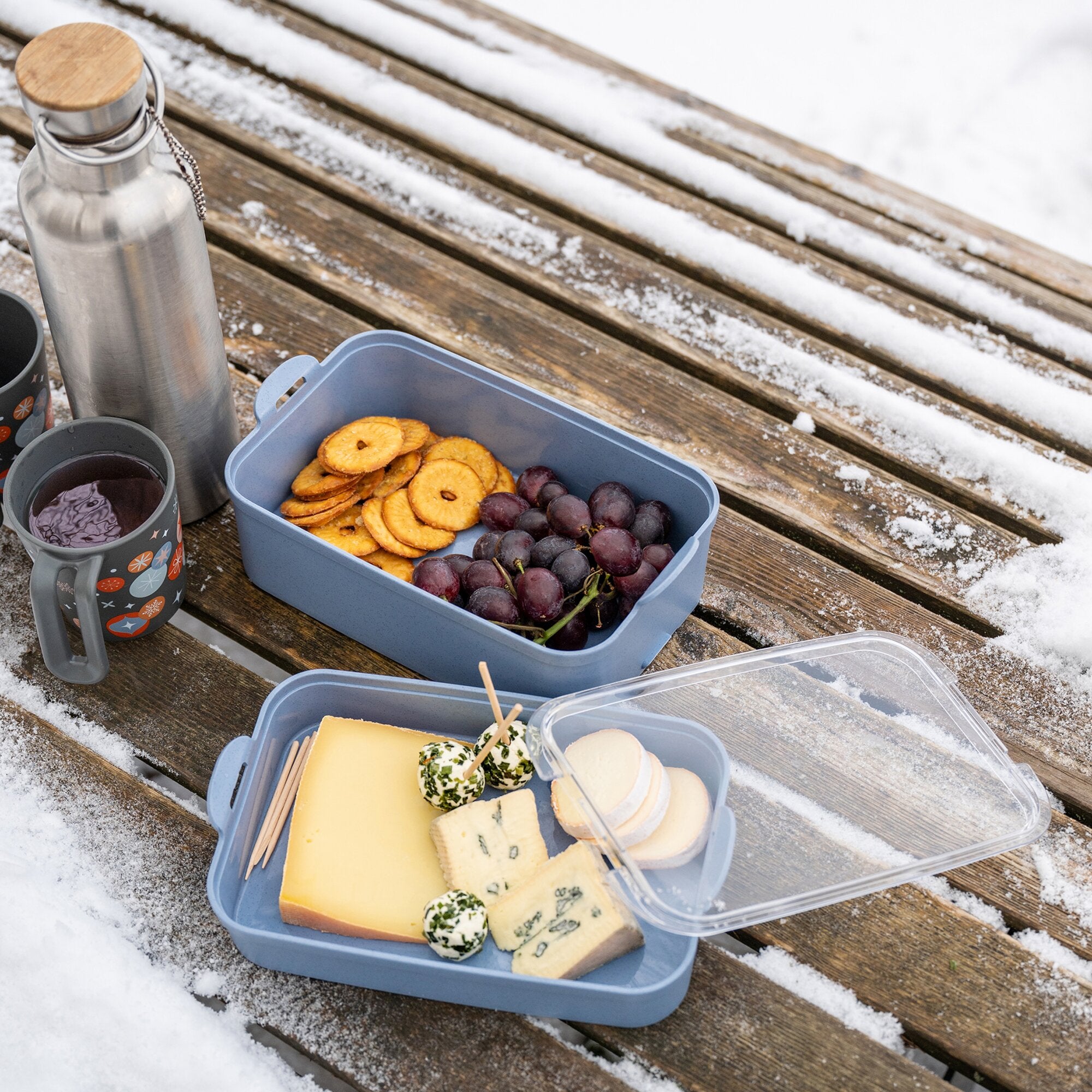 Offene blaue STAKK BENTOBOX 2 Lunchbox mit Crackern und Trauben liegt auf verschneitem Holztisch, daneben Flasche aus Edelstahl mit Holzdeckel
