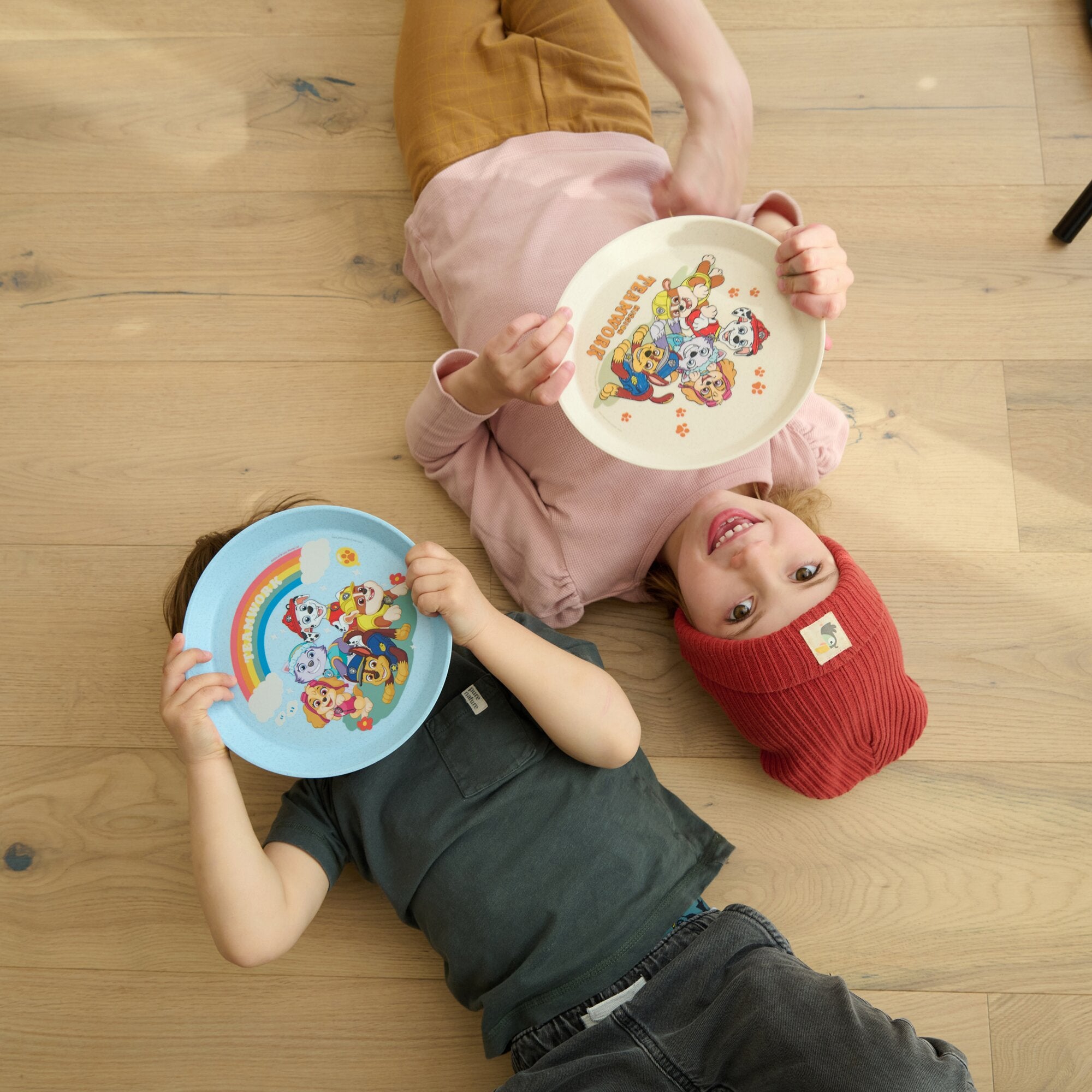 Mehrere Kinder liegen auf Holzboden und halten Paw-Patrol-Teller, weißer Teller mit Paw-Patrol-Charakteren, blauer Teller