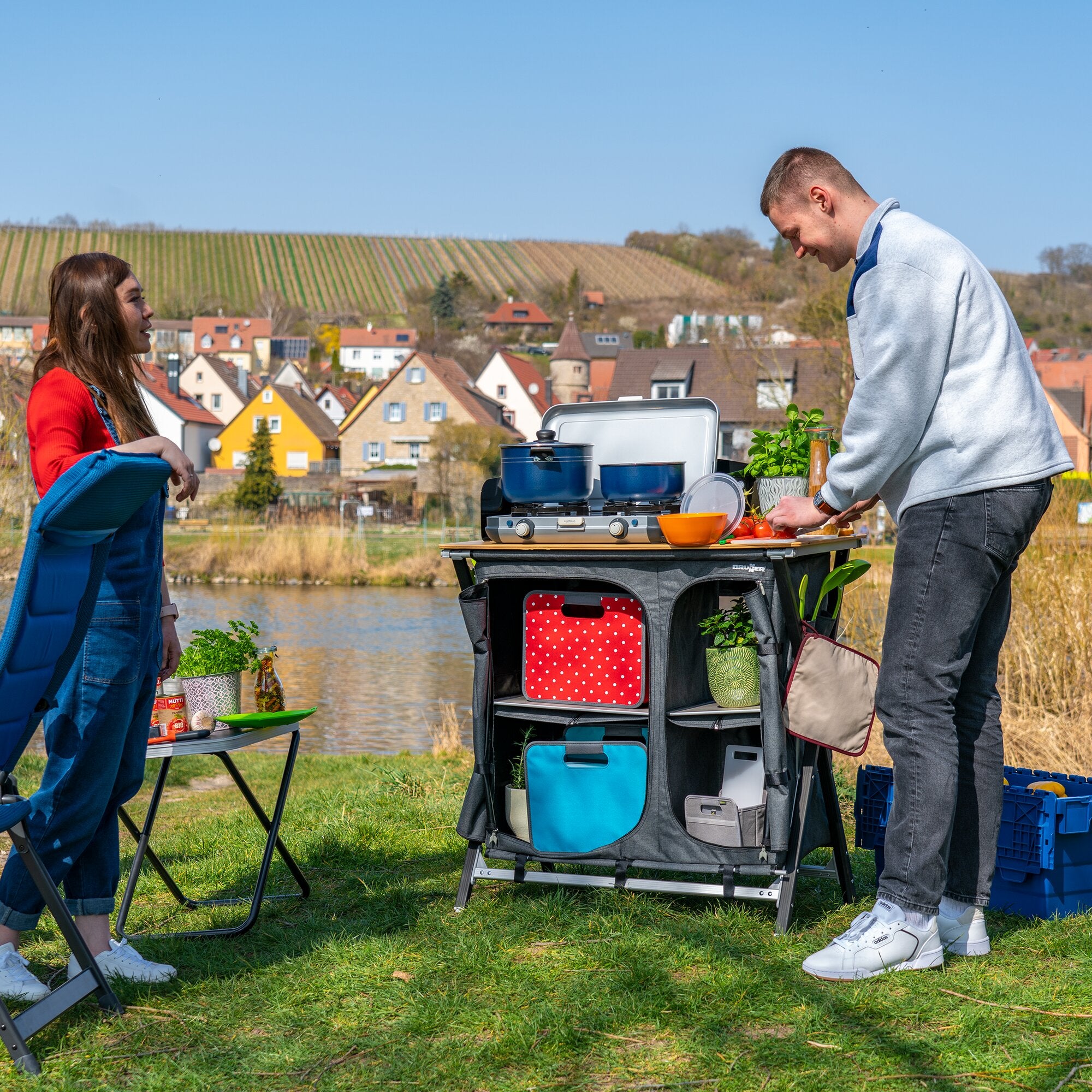Graue Mercury Cross Square Outdoor-Küchenstation mit vielen Taschen; blaue Töpfe und Utensilien stehen bereit, zwei Personen kochen im Freien