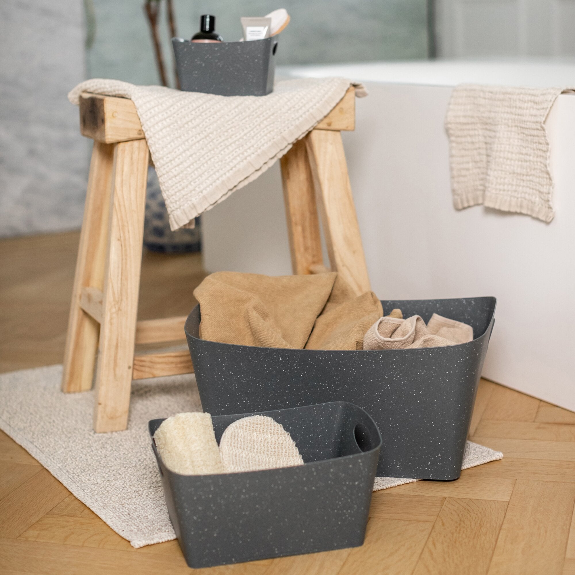 BOXXX M Aufbewahrungsbox grau mit gesprenkelter Oberfläche auf Holzhocker, beige gestrickte Tücher darüber; im Innenraum eine Flasche und eine Tube sichtbar