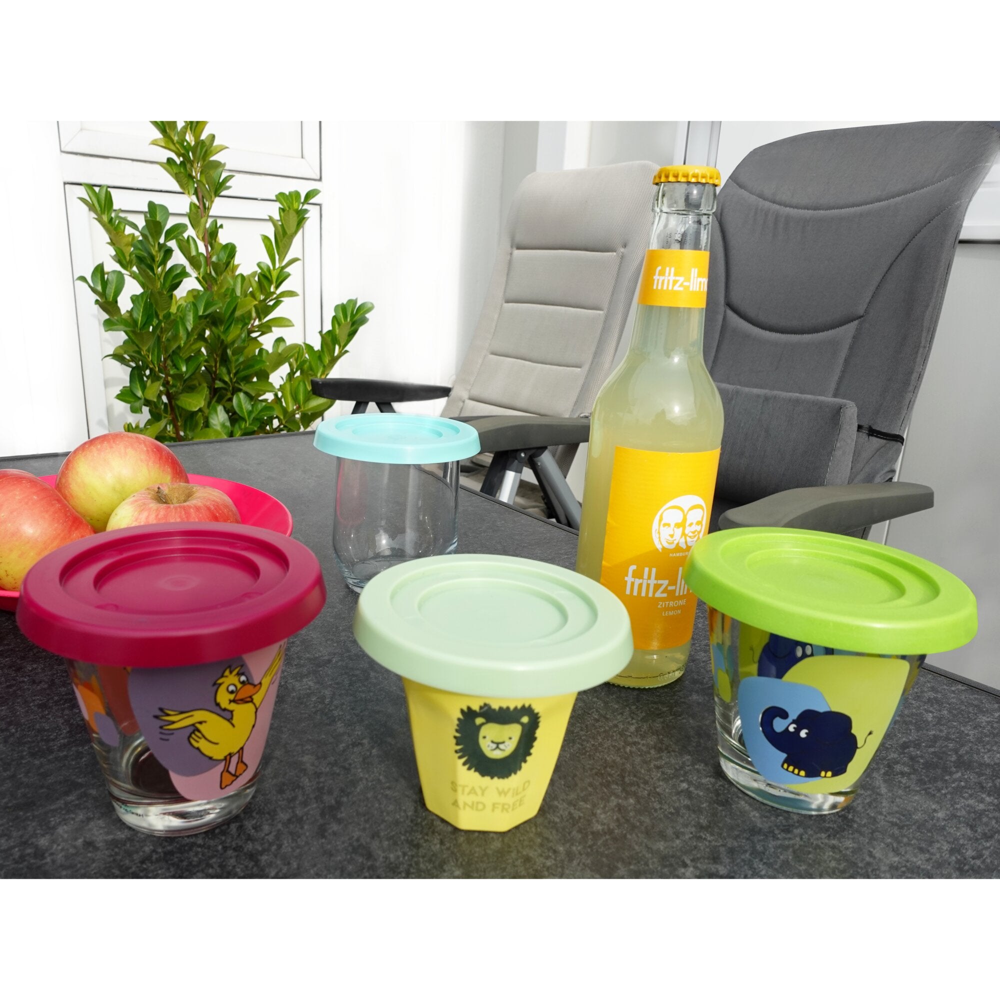 Mehrfarbige Trinkbecher mit bunten Deckeln stehen auf dem Tisch; roter, grüner und blauer Deckel mit Tiermotiven, Obst und Pflanze im Hintergrund