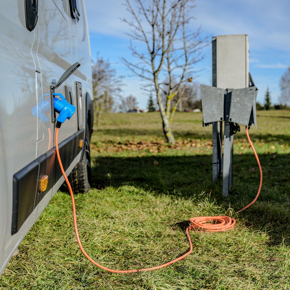 Oranges Camping-Verlängerungskabel mit blauem CEE-Stecker am Wohnmobil angeschlossen, Kabel zieht über Gras und liegt aufgewickelt auf dem Boden