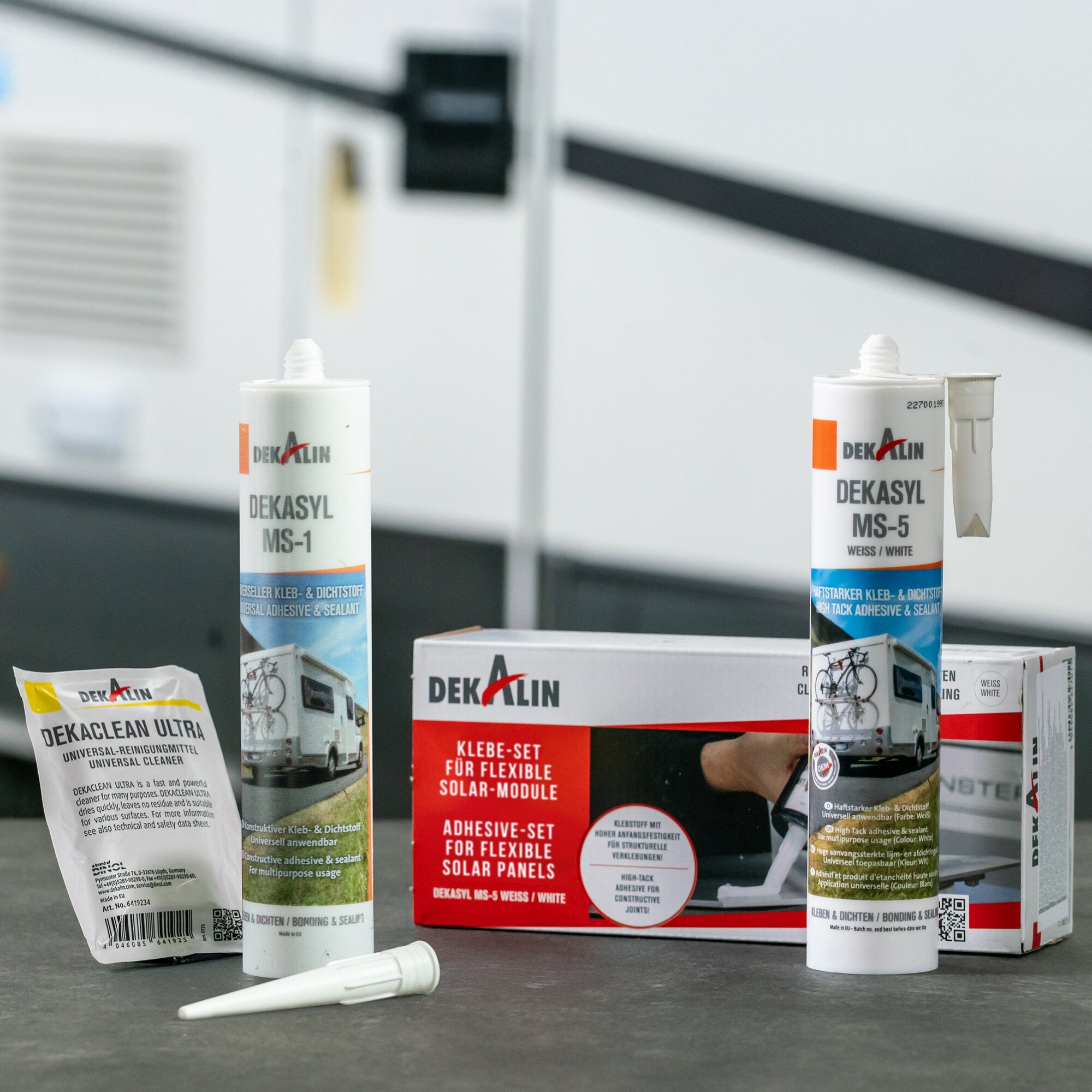 Mehrere weiße Klebstofftuben mit Düsen stehen auf dem Boden, im Hintergrund Kartonverpackungen mit rotem Schild KLEBE-SET FÜR FLEXIBLE SOLAR MODULES und DEKASYL MS-1/MS-5 Verpackungen