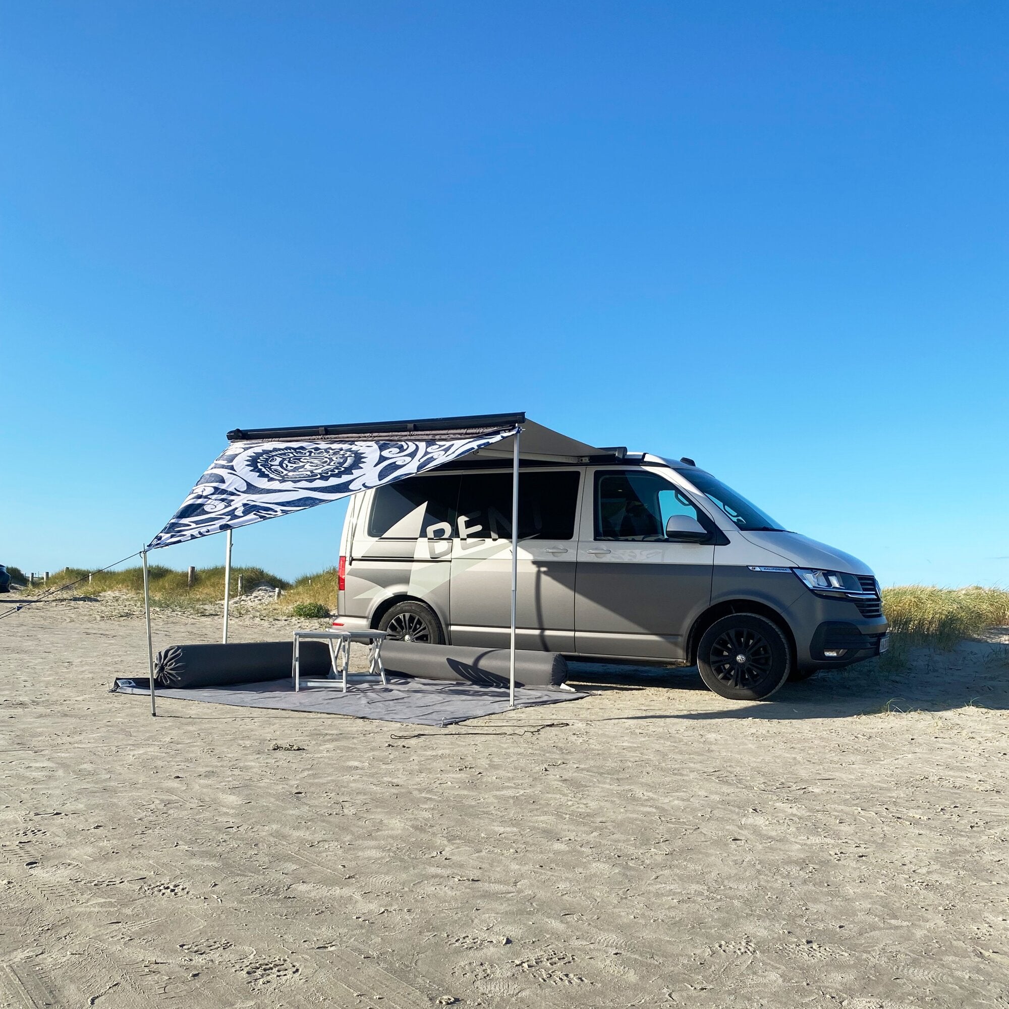 Sonnensegel Zip Canvas New Zealand mit schwarz-weißem Muster ist am Camper-Van am Strand gespannt, daneben Tisch und Matte