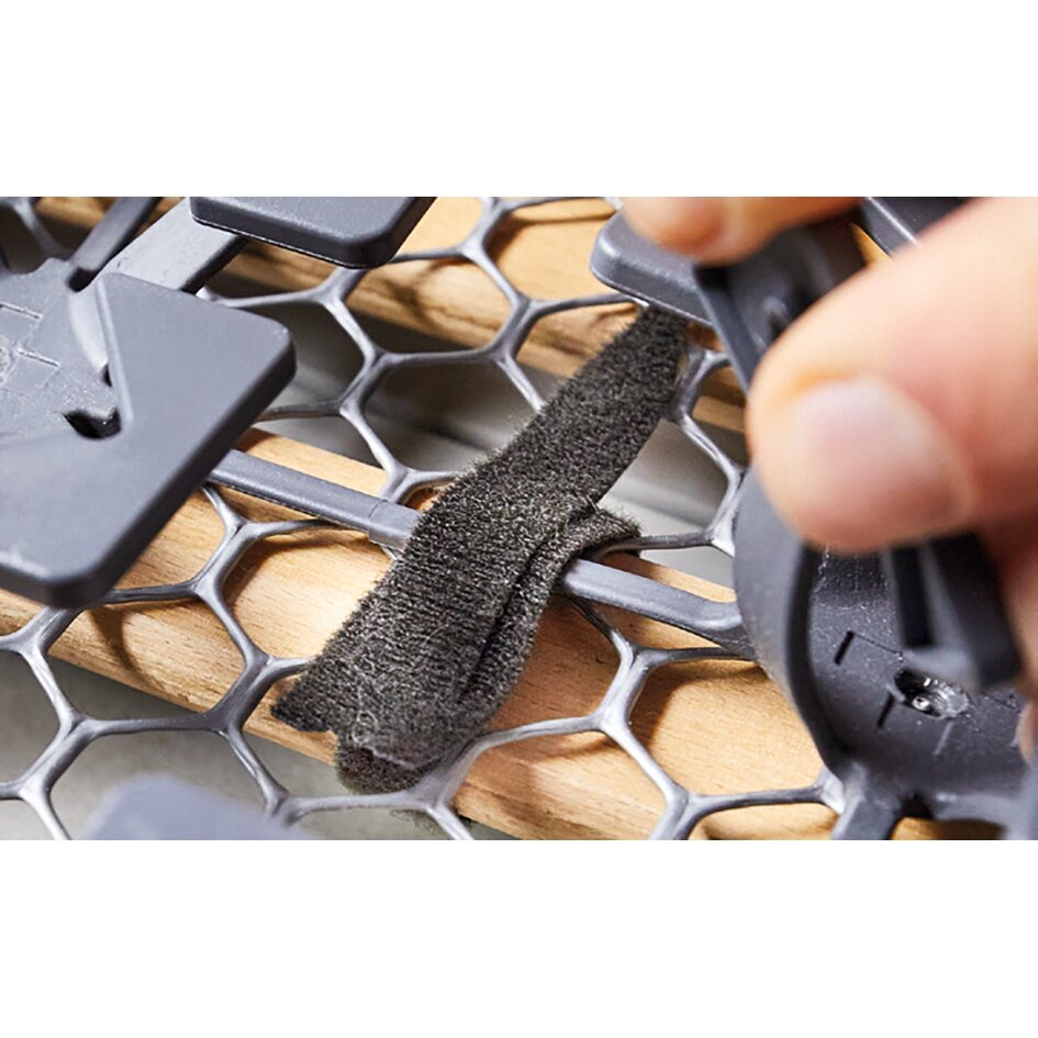 Detailaufnahme eines hexagonal geformten Kunststoffgitters; eine Hand befestigt eine schwarze Klettschlaufe am Rand des Froli Schlafset für VW T5/T6 California