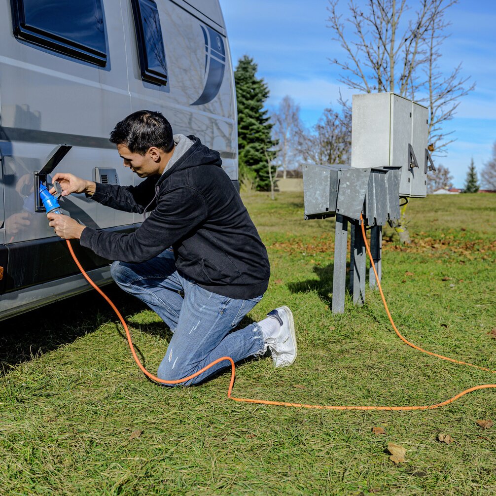 Ein Mann kniet im Gras neben einem Wohnmobil und verbindet eine orange CEE-Verlängerung mit einem blauen Stecker zu den Außenverteilerkästen