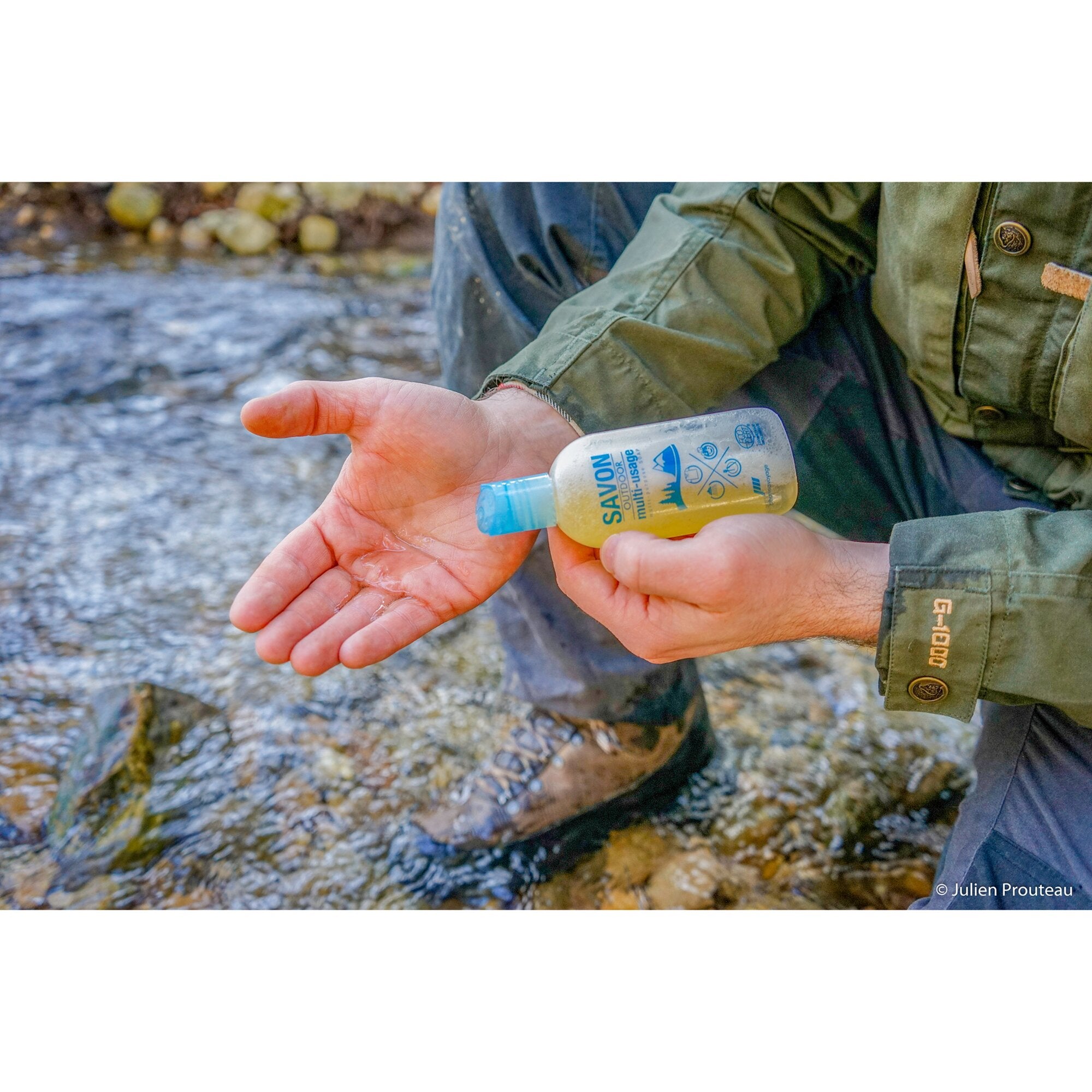Person in einer olivgrünen Outdoor-Jacke hält eine kleine Flasche Savon Outdoor multi-usage Seife mit blauem Deckel; Etikett sichtbar, Flussufer im Hintergrund