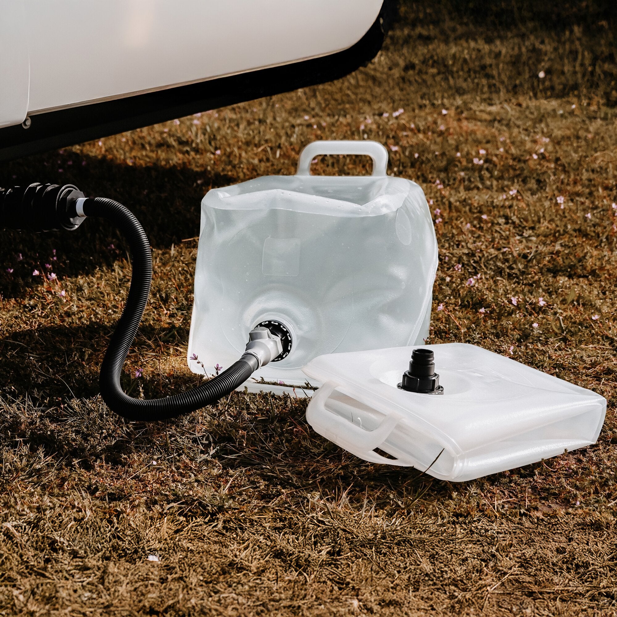 Grauwasser-Sammelsystem 2 x 20 l, transparenter Tank mit Griff und Schlauchanschluss, im Gras neben weiteren weißen Behälterteilen