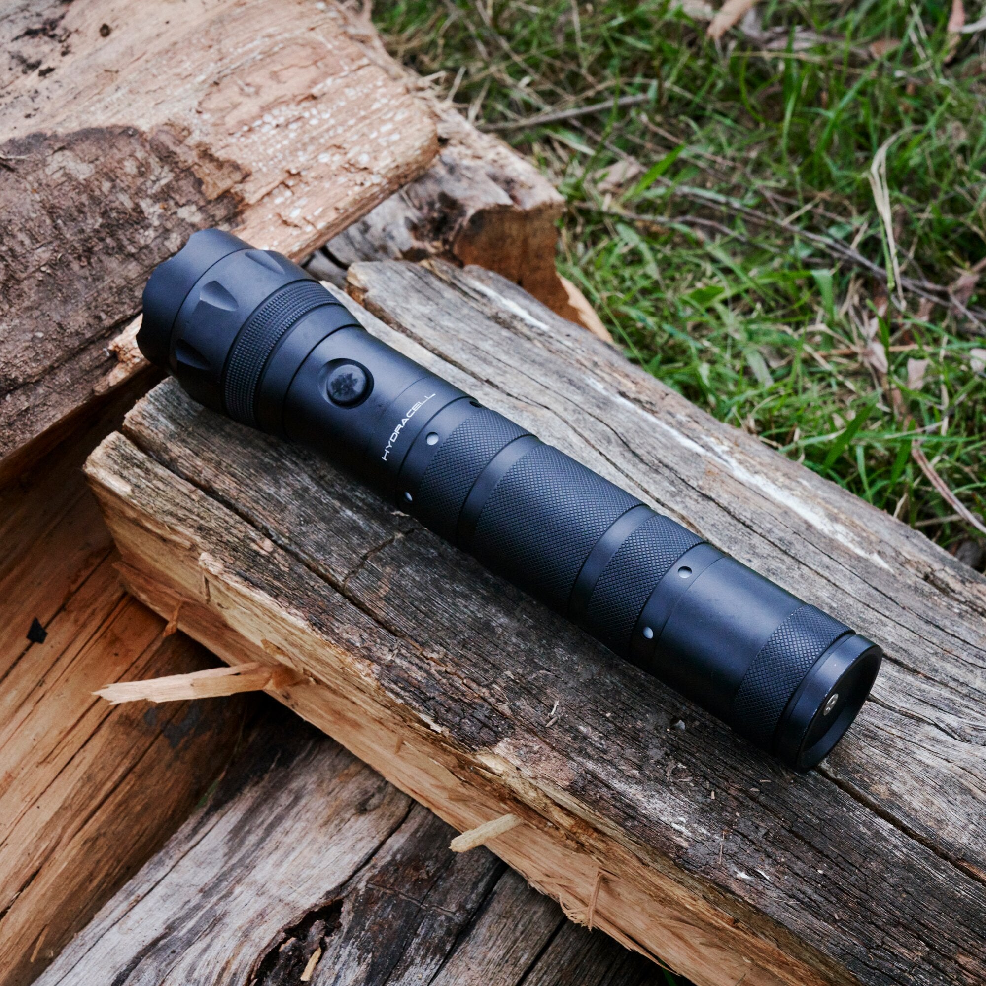 Schwarze, matte Taschenlampe mit rutschfestem, genopptem Griff liegt schräg auf einem Holzstamm, Gras im Hintergrund