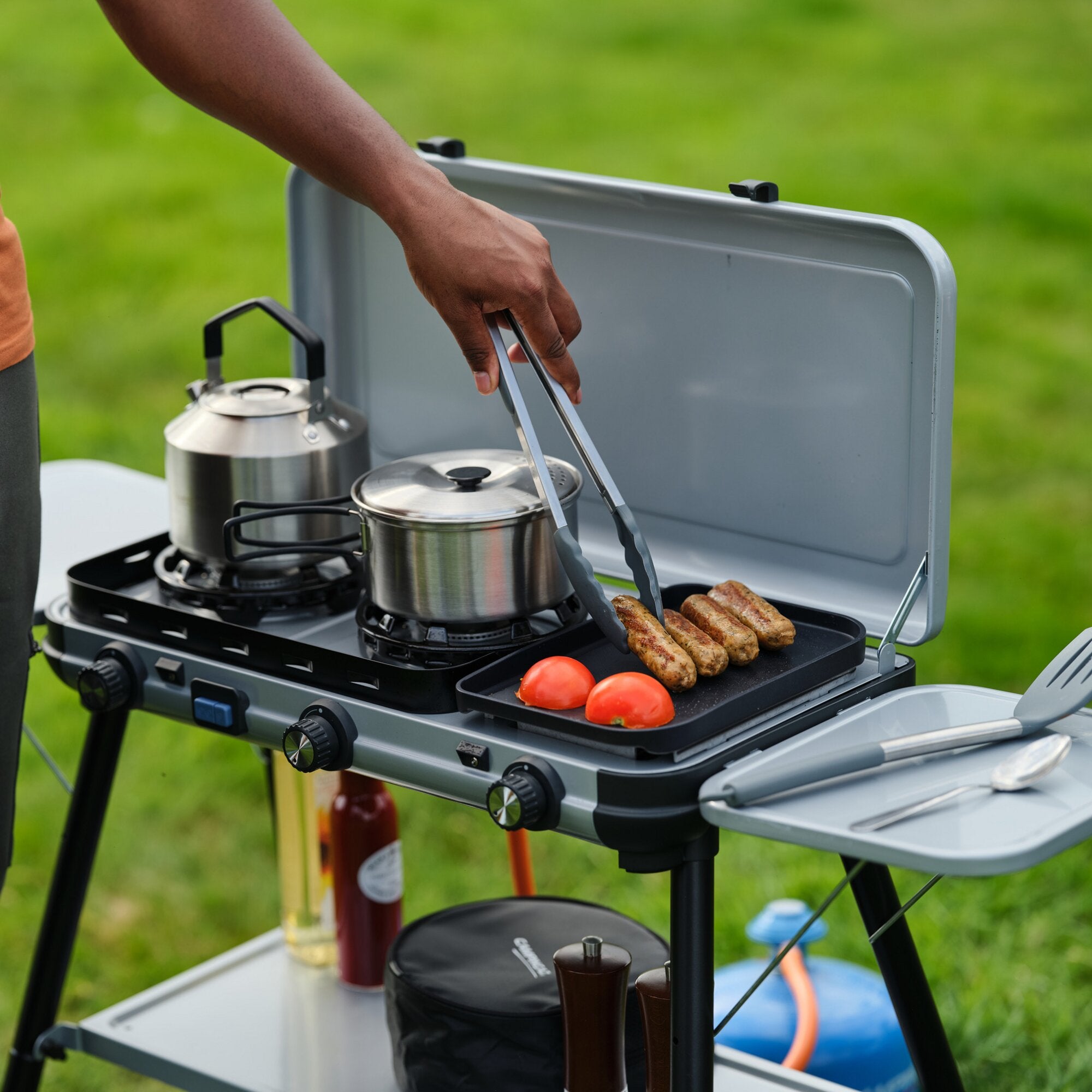 Gaskocher Camping Kitchen 2 Multi-Cook Plus im Freien; zwei Brenner, zwei Töpfe, Grillplatte mit Würstchen und Tomaten, Person bedient