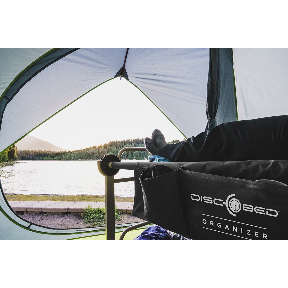 Zeltinnenblick mit Feldbett; seitliche schwarze Seitentasche am Bettgestell trägt Disc-Bed-Organizer-Logo; Person ruht mit Füßen an der Bettkante, See