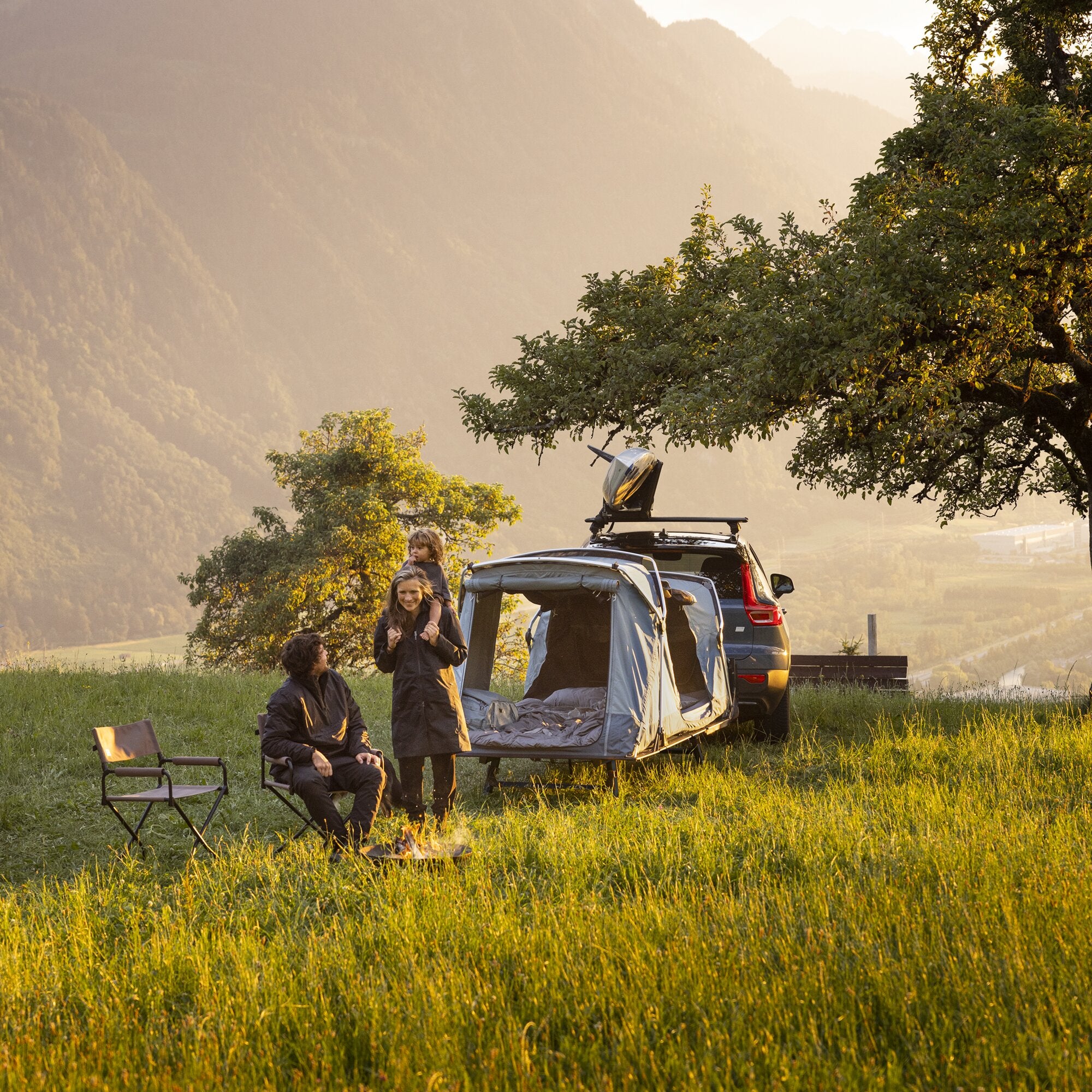Thule Outset Anhängerkupplungszelt am Heck eines SUVs geöffnet, Familie sitzt vor dem Wagen, Campingstuhl, Lagerfeuer, und im Hintergrund Berglandschaft