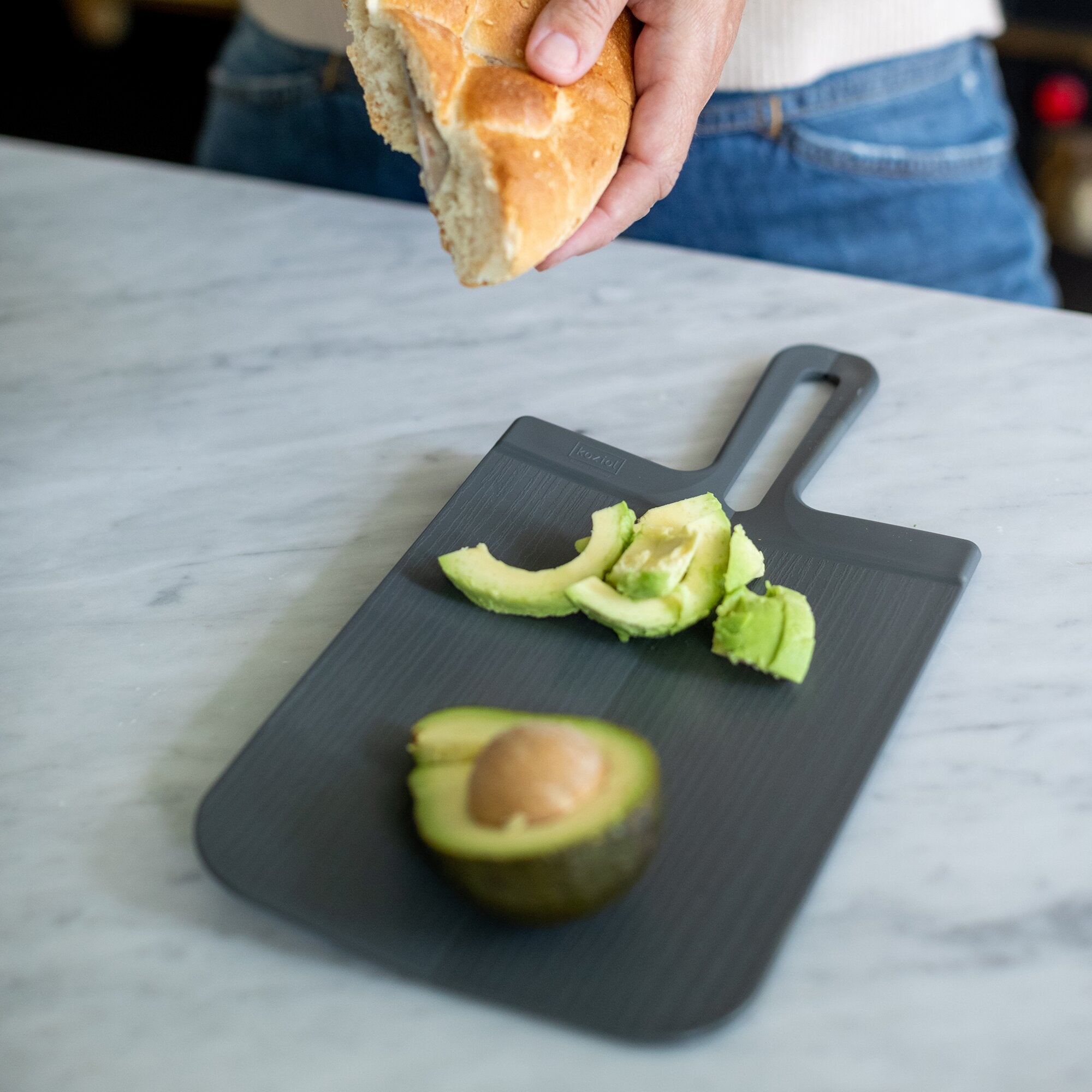 Graues Schneidebrett SNAP S auf heller Arbeitsfläche, Avocadostücke darauf, während eine Hand darüber ein Brot hält