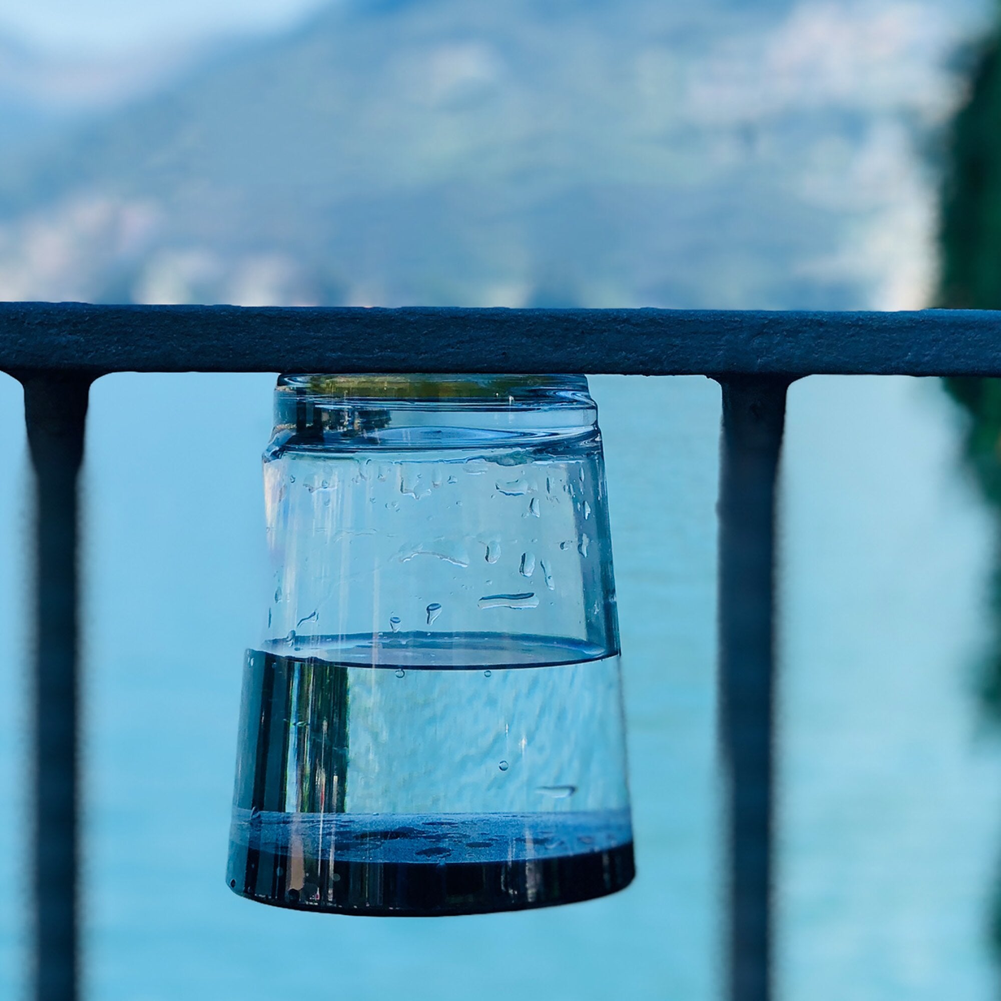 Nahaufnahme eines transparenten Glastrinkbechers mit Wassertropfen an der Außenseite, vor dunkler Metallstange und unscharf blauem Hintergrund