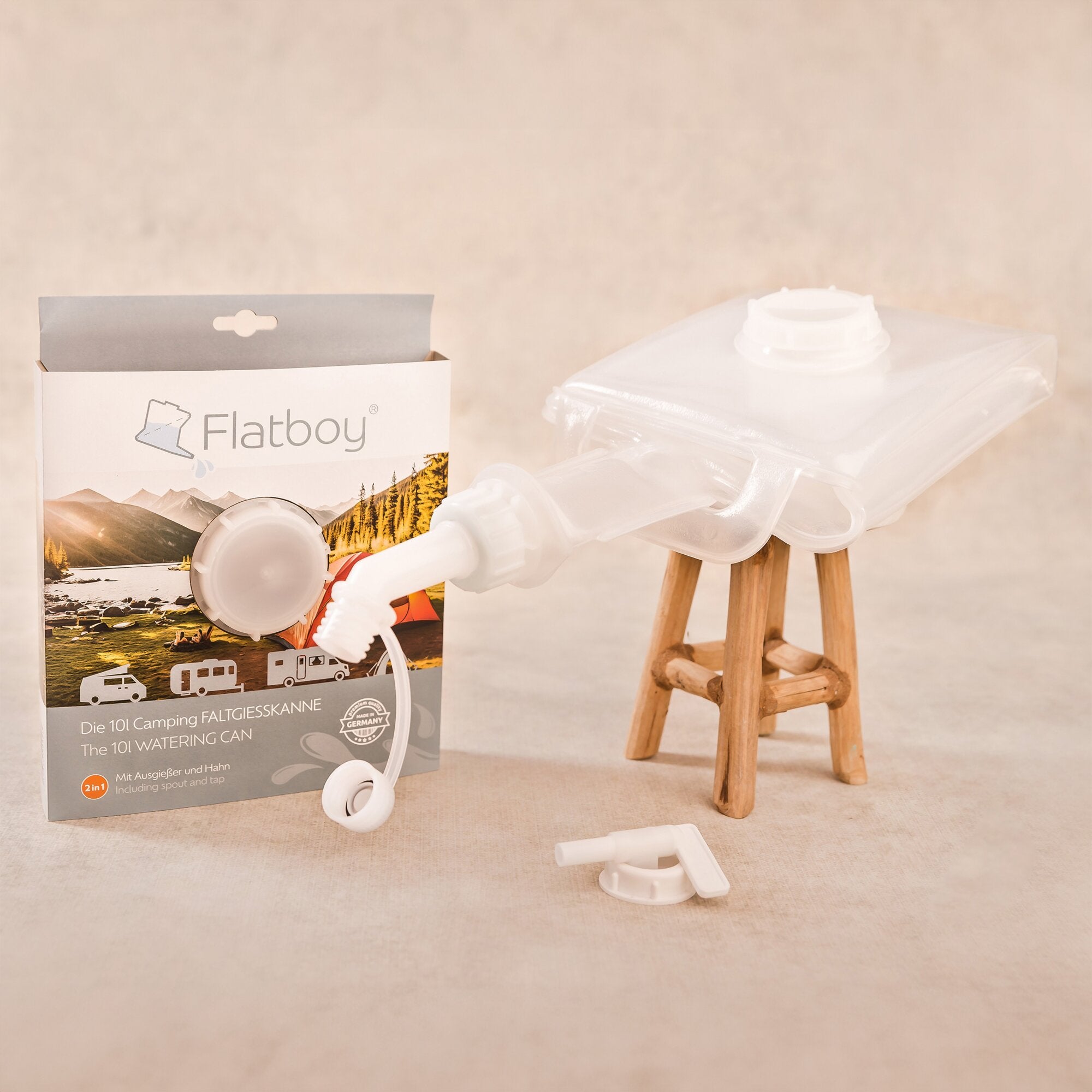 Verpackung der faltbaren Gießkanne Flatboy 10 l steht neben losem weißen Kunststoffzubehör, vor beigem Hintergrund und einem Holzhocker