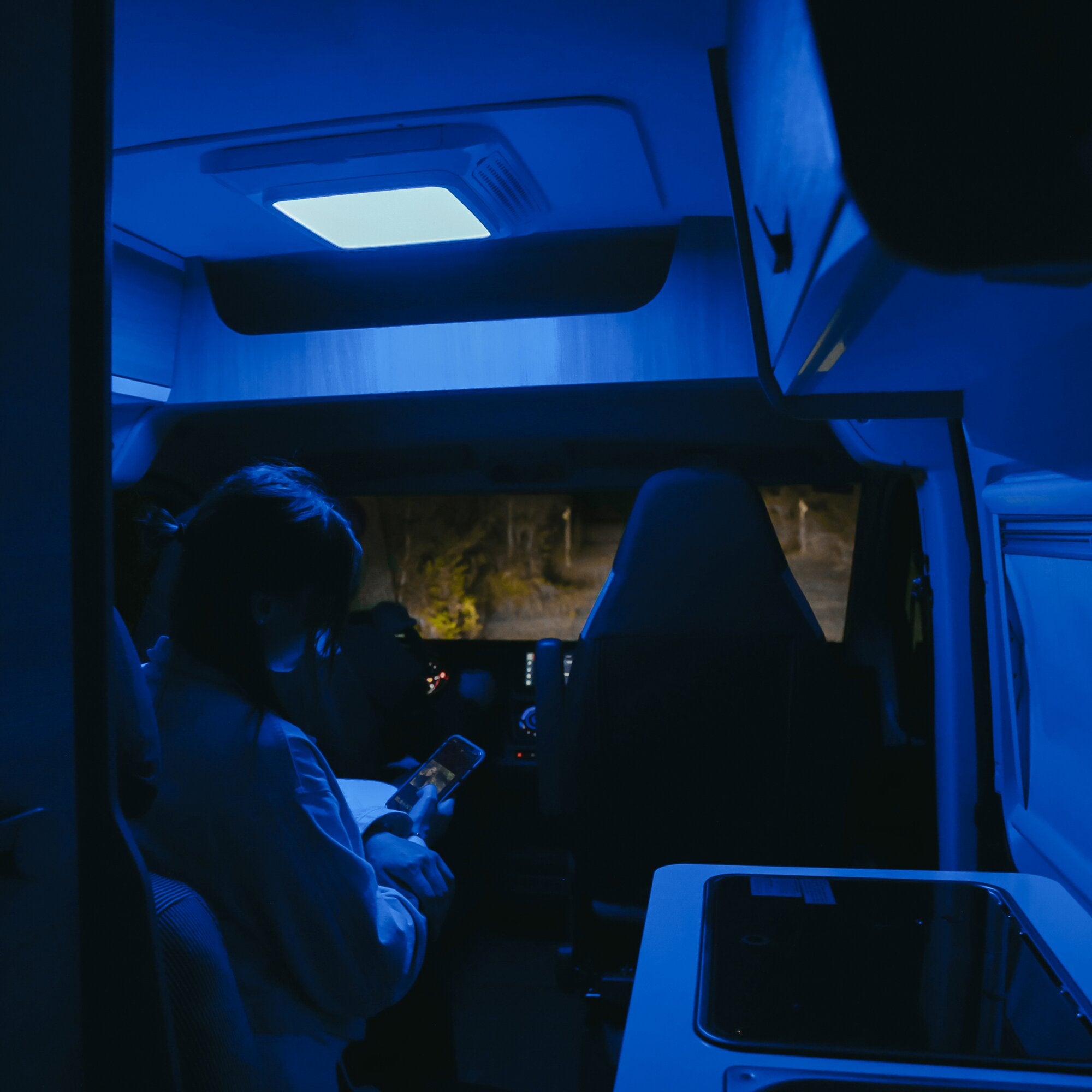 Innenraum eines Campers bei Nacht, blau beleuchtet; eine rechteckige Deckenleuchte Luftverteiler Lightbox für Klimaanlage ist sichtbar, umgeben von Sitzbank und Fahrerraum