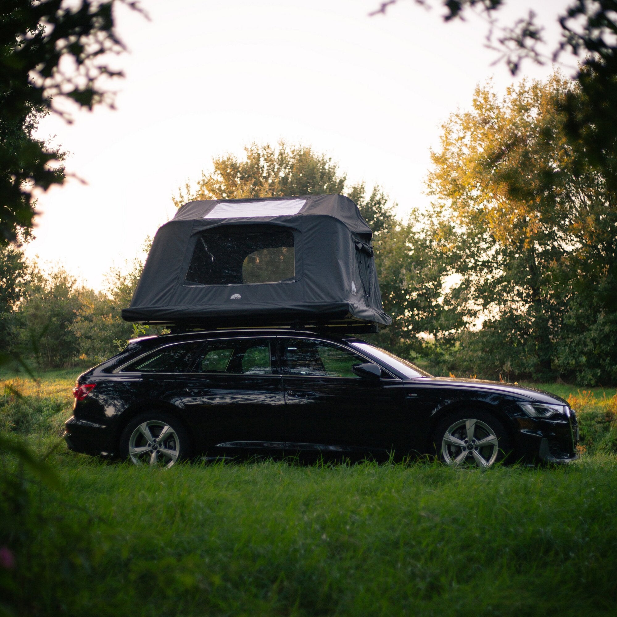 Schwarzer SUV mit einem dunkelgrauen Dachzelt Roof Space 4 auf dem Dach, Fensterfront sichtbar, umgeben von Wald und Gras