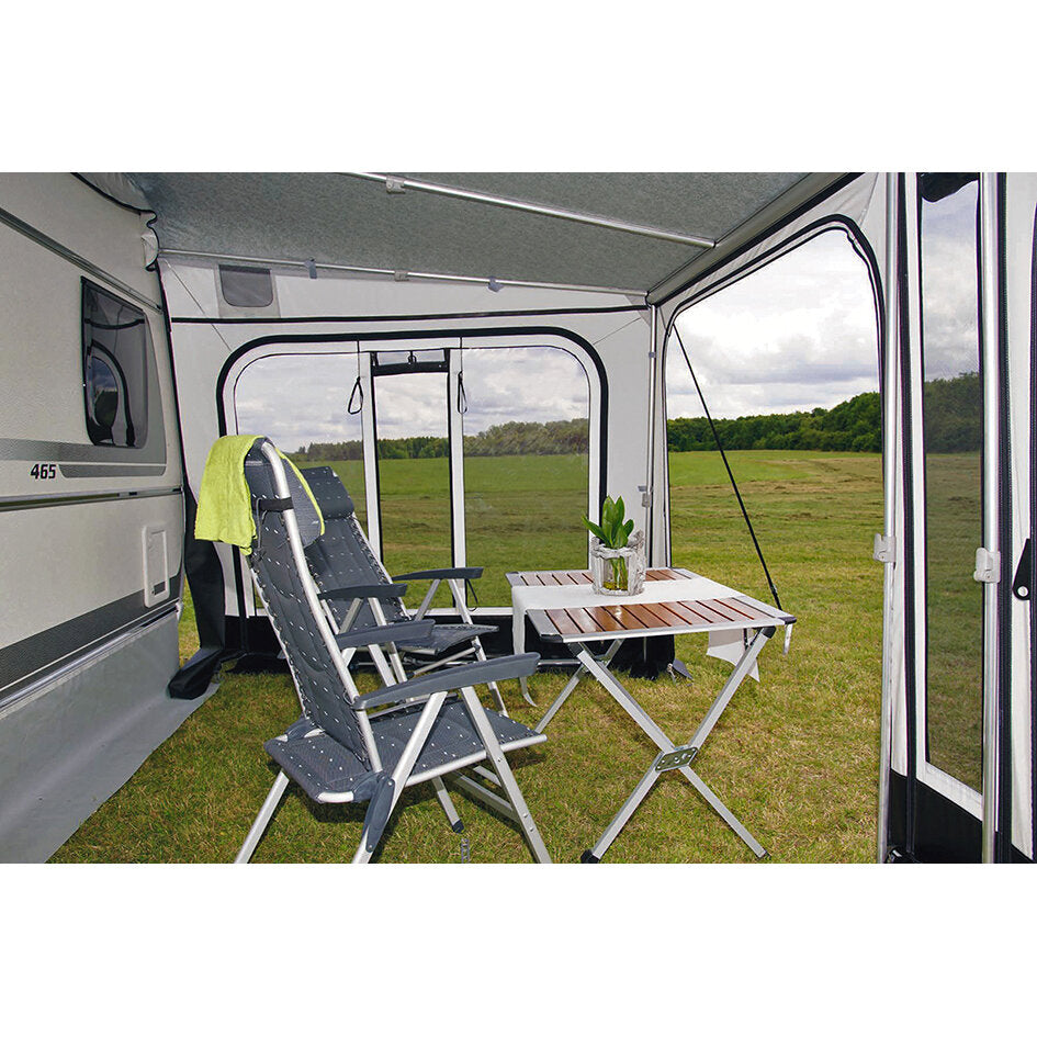 Innenraum einer Caravan-Markisen-Seitenwand mit zwei Klappstühlen, kleinem Klapptisch, Pflanze und grünem Handtuch; Blick nach draußen auf eine Wiese