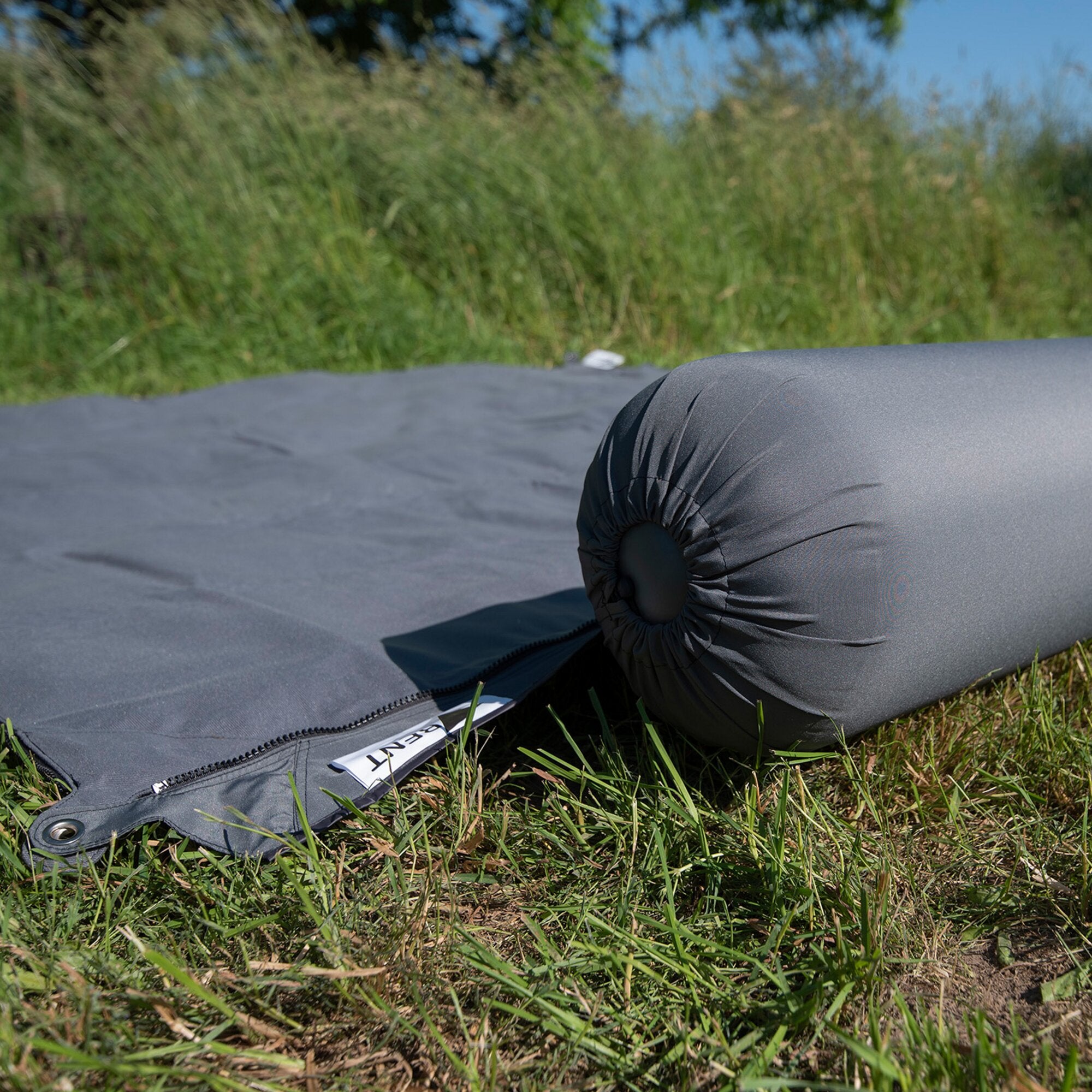 Dunkelgraue Kissenrolle Zip XL Lounger mit Reißverschluss liegt quer auf einer grauen Matte im Freien, Gras und blauer Himmel im Hintergrund