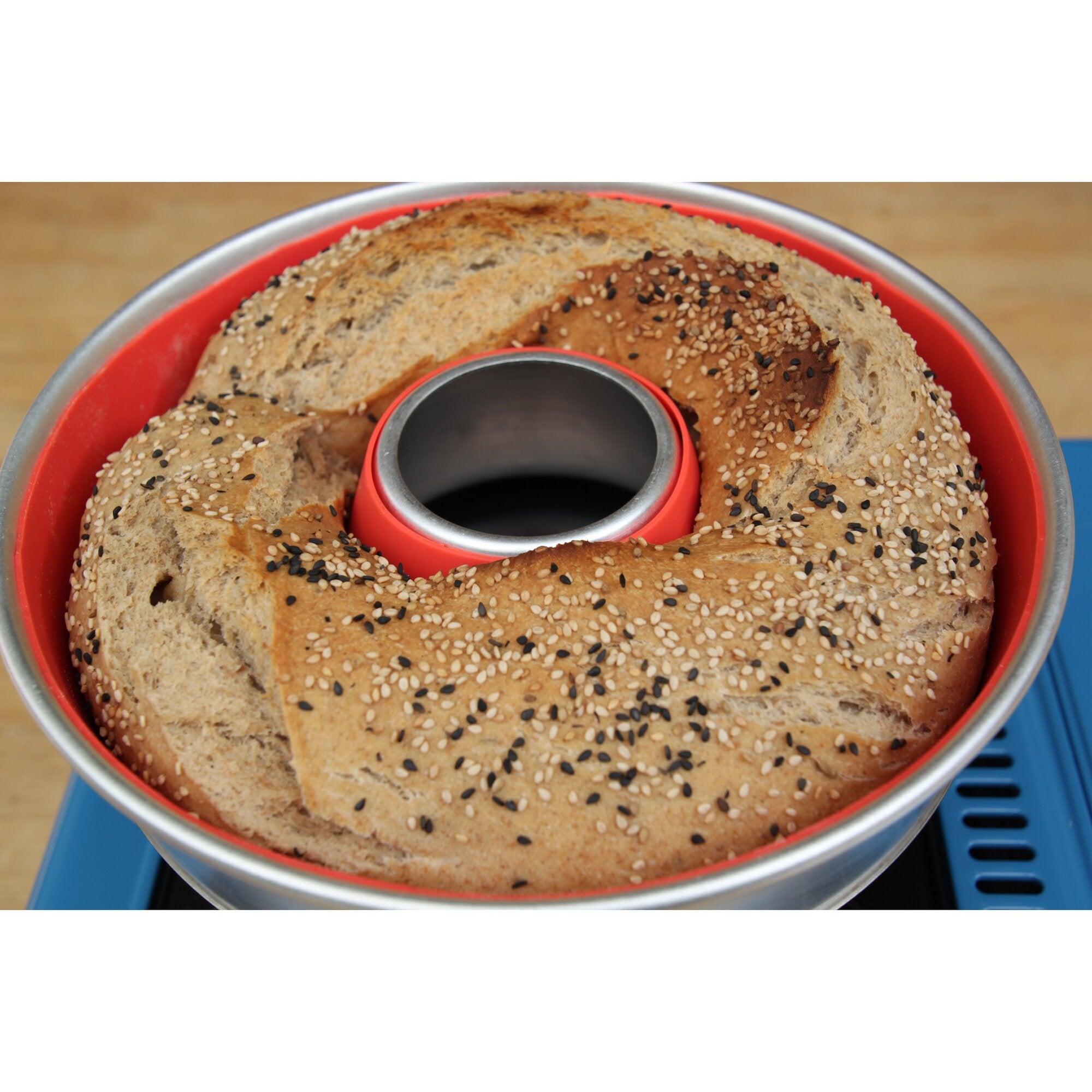 Runde rote Silikonbackform mit zentralem Metallring, darin liegt ein Kuchenring mit Sesamsamen bestreut, Hintergrund Holzplatte