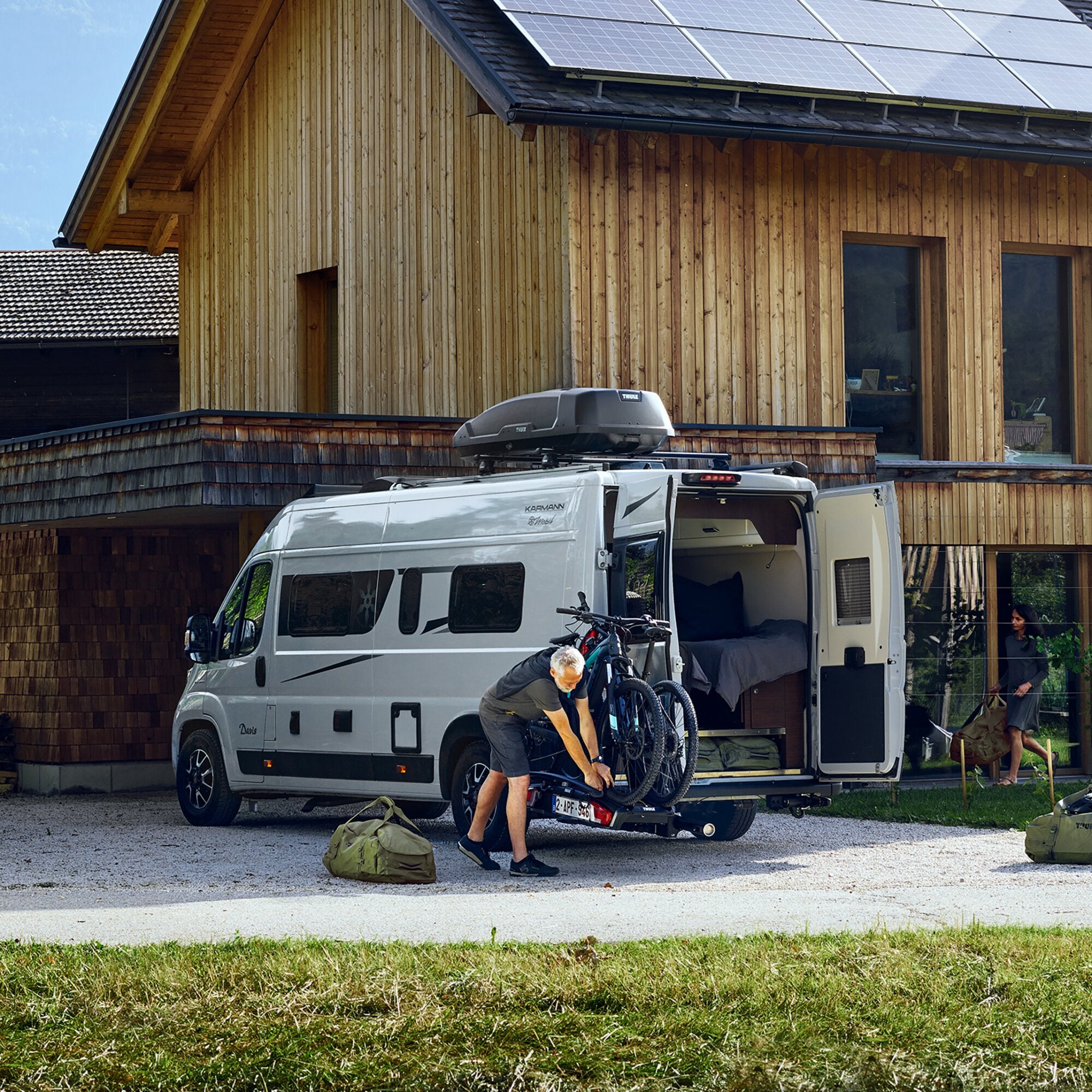 Wohnmobil mit Heck-Fahrradträger, zwei Fahrräder montiert, Person beim Beladen, Holzhaus mit Solarmodulen im Hintergrund