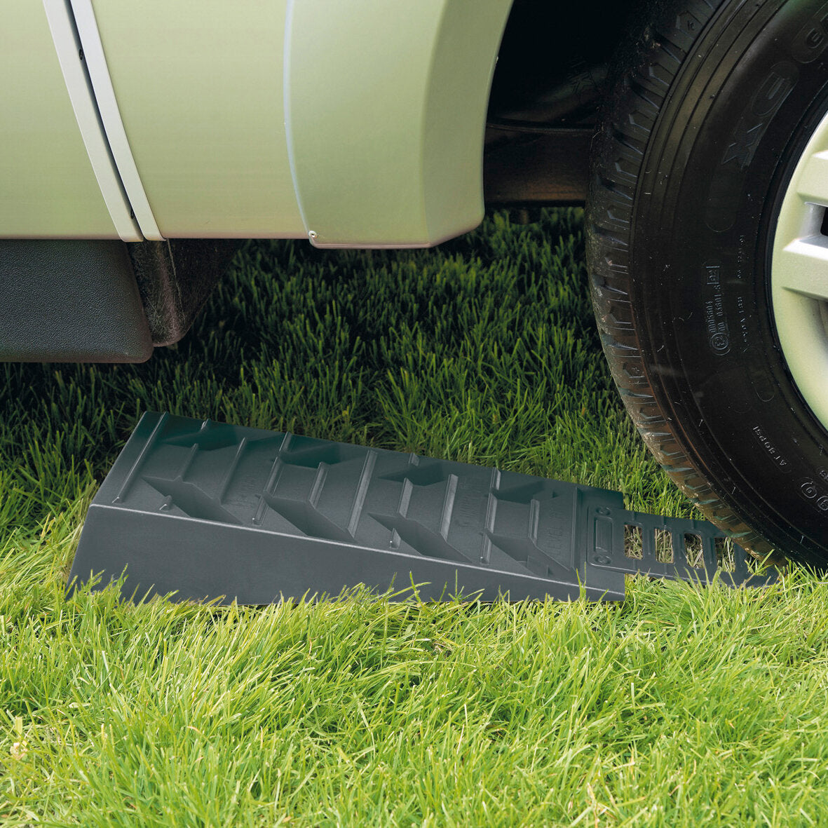 Grauer Kunststoff-Auffahrkeil aus dem Level Pro 2er-Set liegt auf Gras neben dem Reifen eines hellgrünen Autos; die Oberseite zeigt Rippen