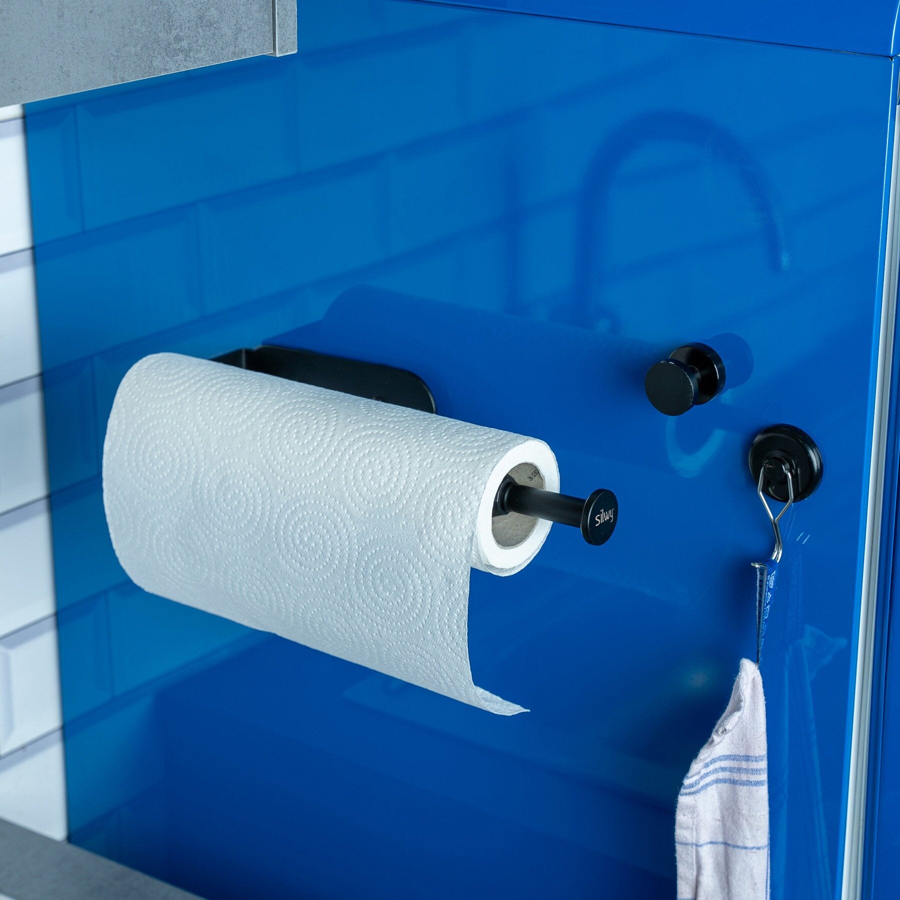 Magnetischer Küchenrollenhalter aus schwarzem Metall mit Papierrolle, an blauer Wand montiert, Endkappen sichtbar, unten hängt ein Tuch